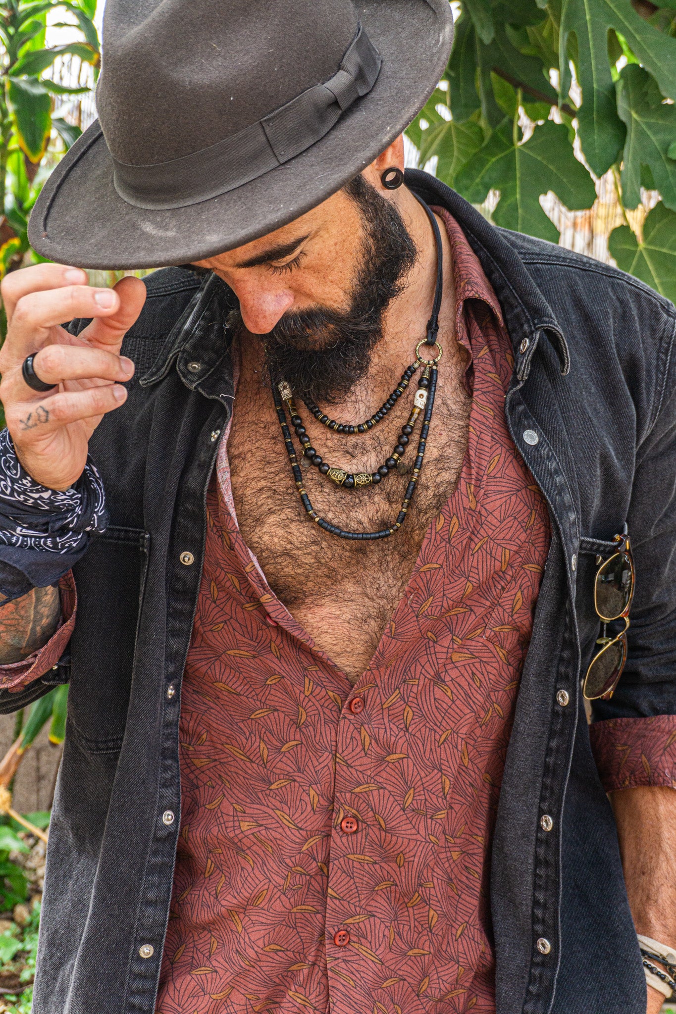 Man wearing a dark hat, black jacket, and patterned shirt with necklaces outdoors.