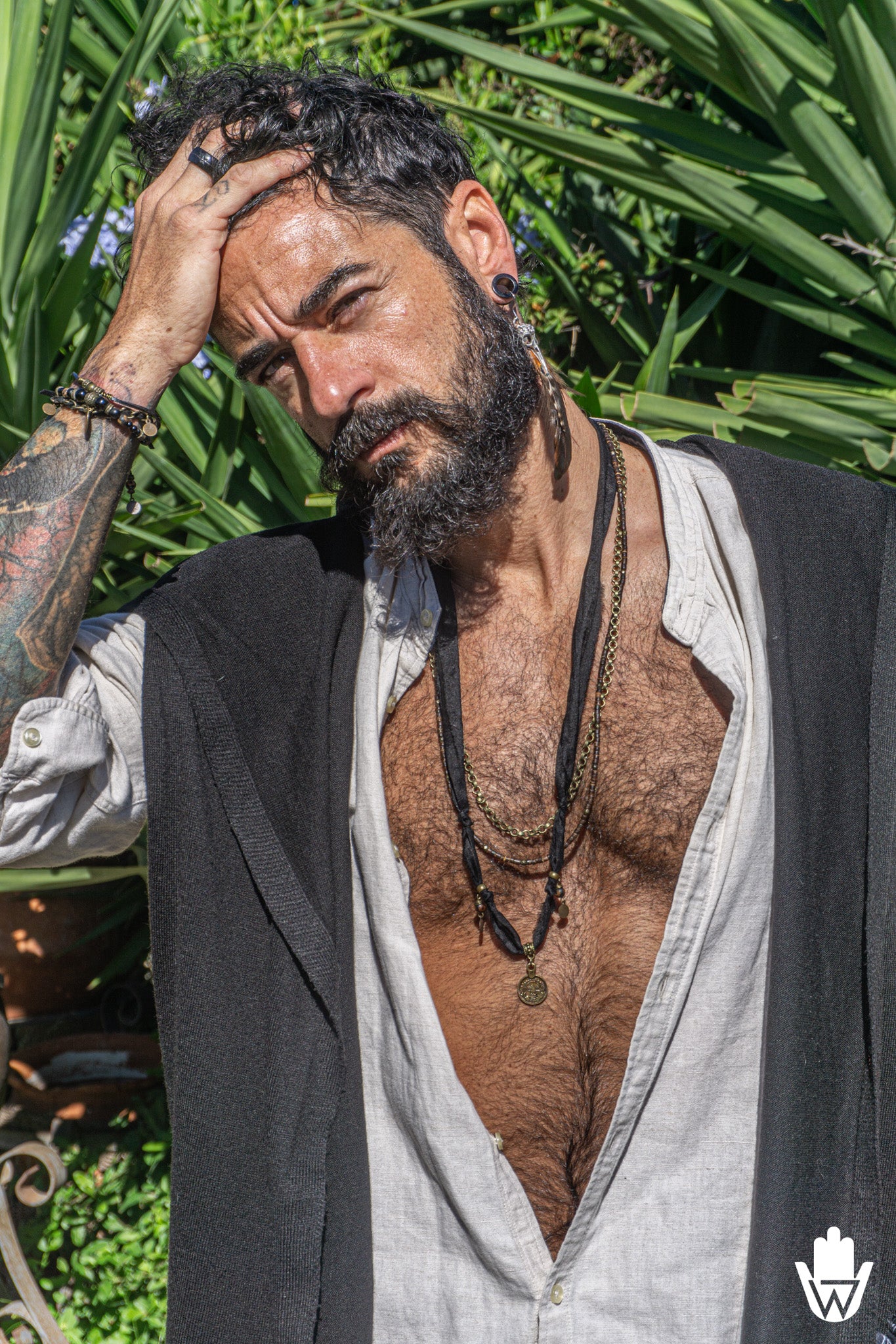 A man with a beard and tattoos poses outdoors in front of green foliage, wearing an open light gray linen shirt, a dark gray vest, layered black necklaces, and a ring.