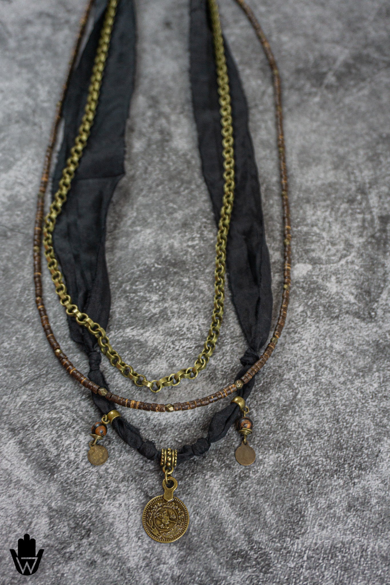A bohemian-style multi-strand necklace featuring a gold-tone chain, brown beaded strands, black fabric ribbons, and bronze coin pendants, displayed on a textured gray background.