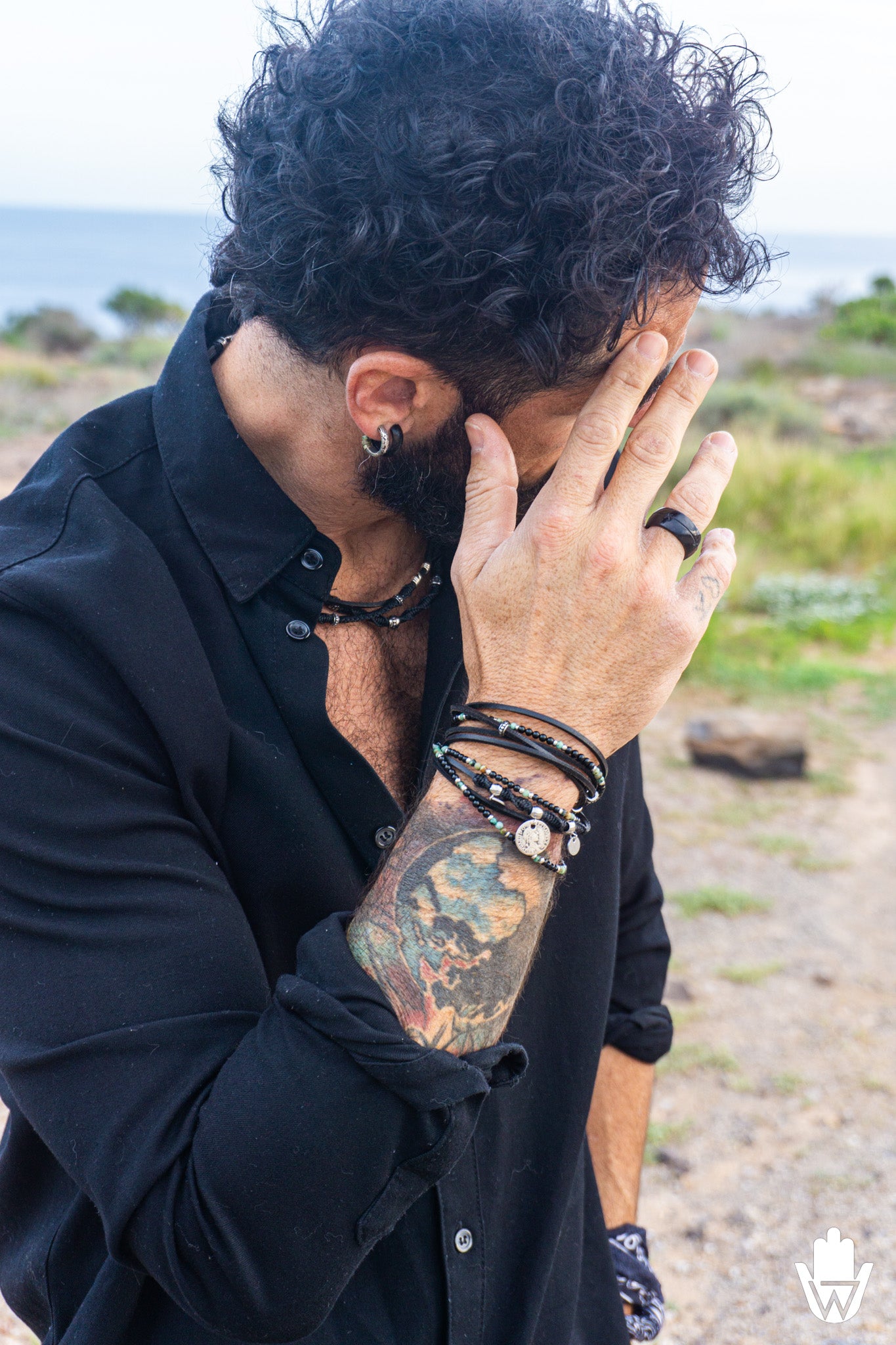 Man with tattoos and boho jewelry covering his face outdoors