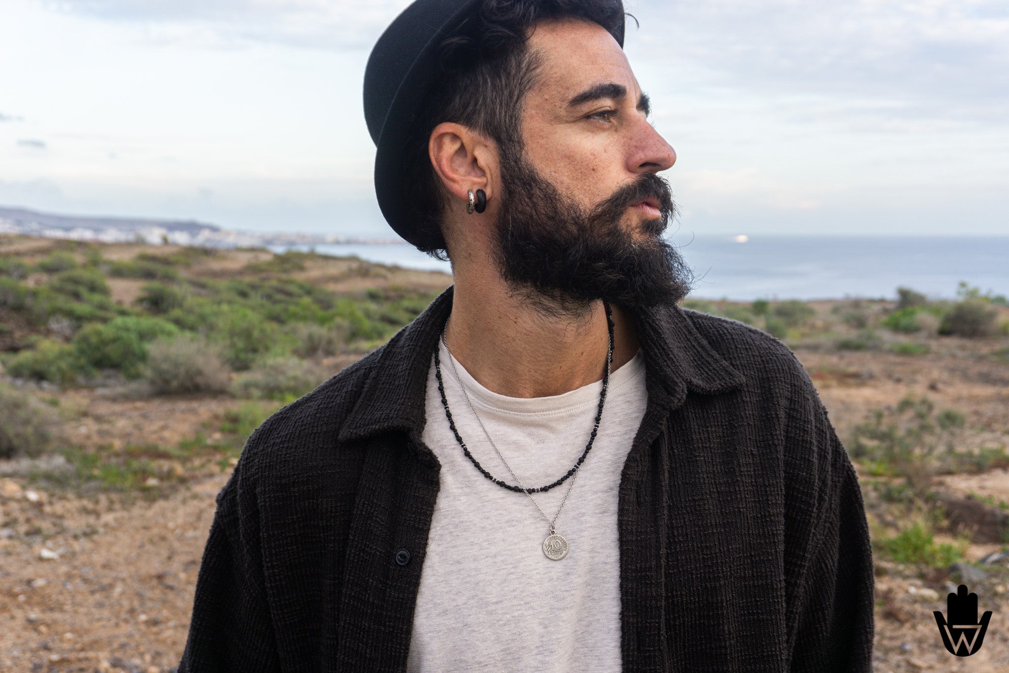 A man with a beard and black hat wearing a black textured overshirt over a light gray t-shirt, standing outdoors in a dry, scrub-covered landscape with the ocean in the background.