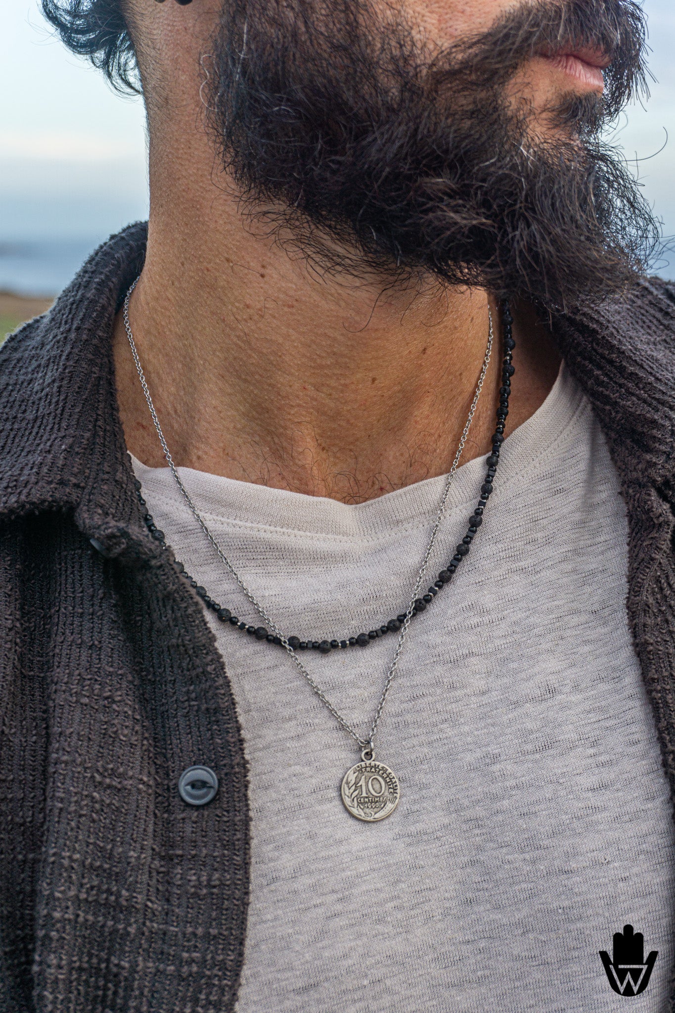 A close-up of a man's neck and chest wearing two layered necklaces: a silver chain with a circular pendant and a black beaded chain. He is wearing a light gray t-shirt and a dark brown button-up shirt, with a blurred outdoor background.