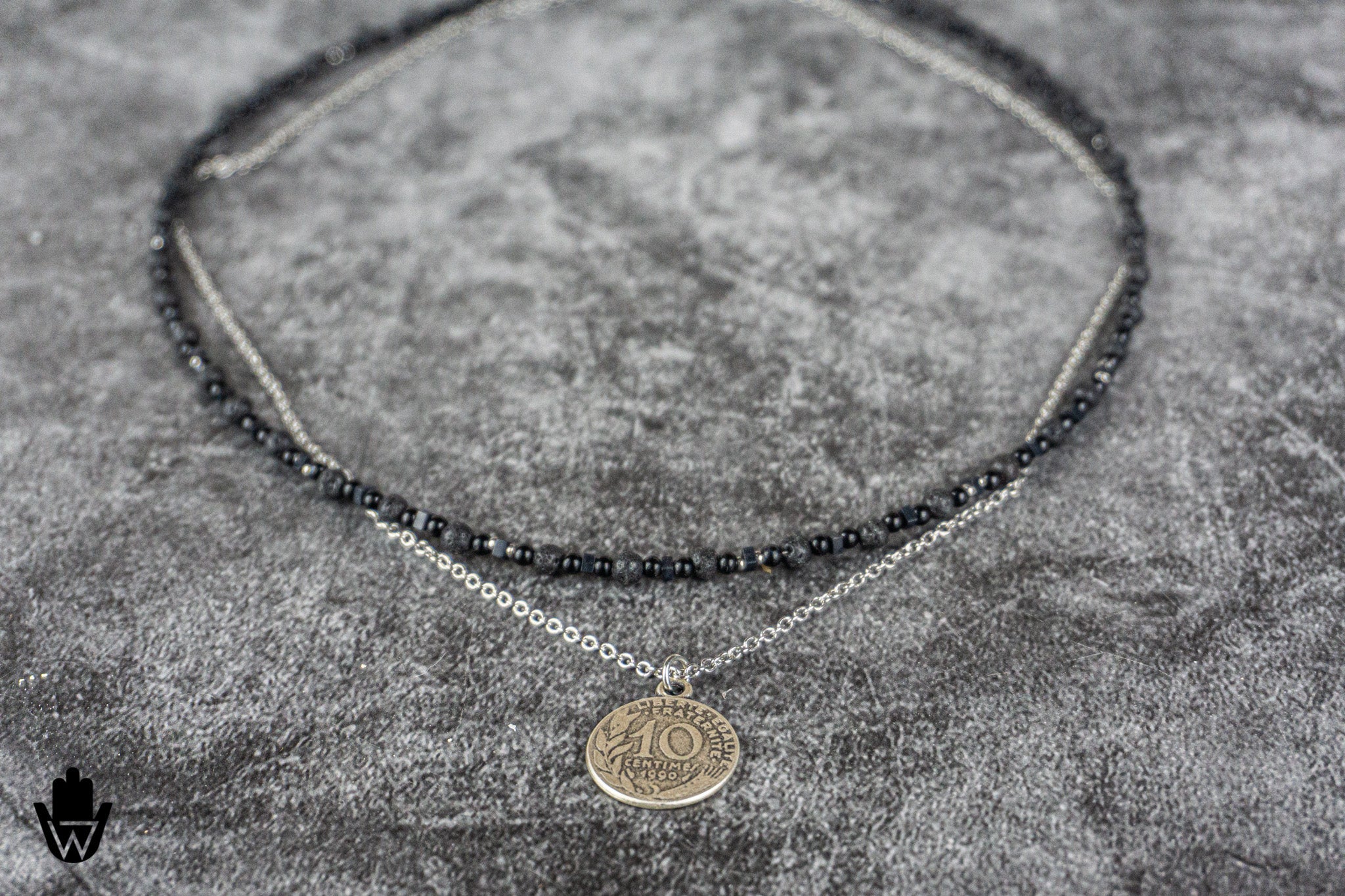 A double-strand necklace featuring a top layer of dark gray faceted beads and a bottom layer of silver chain with a round gold-toned coin pendant, displayed on a textured gray background.