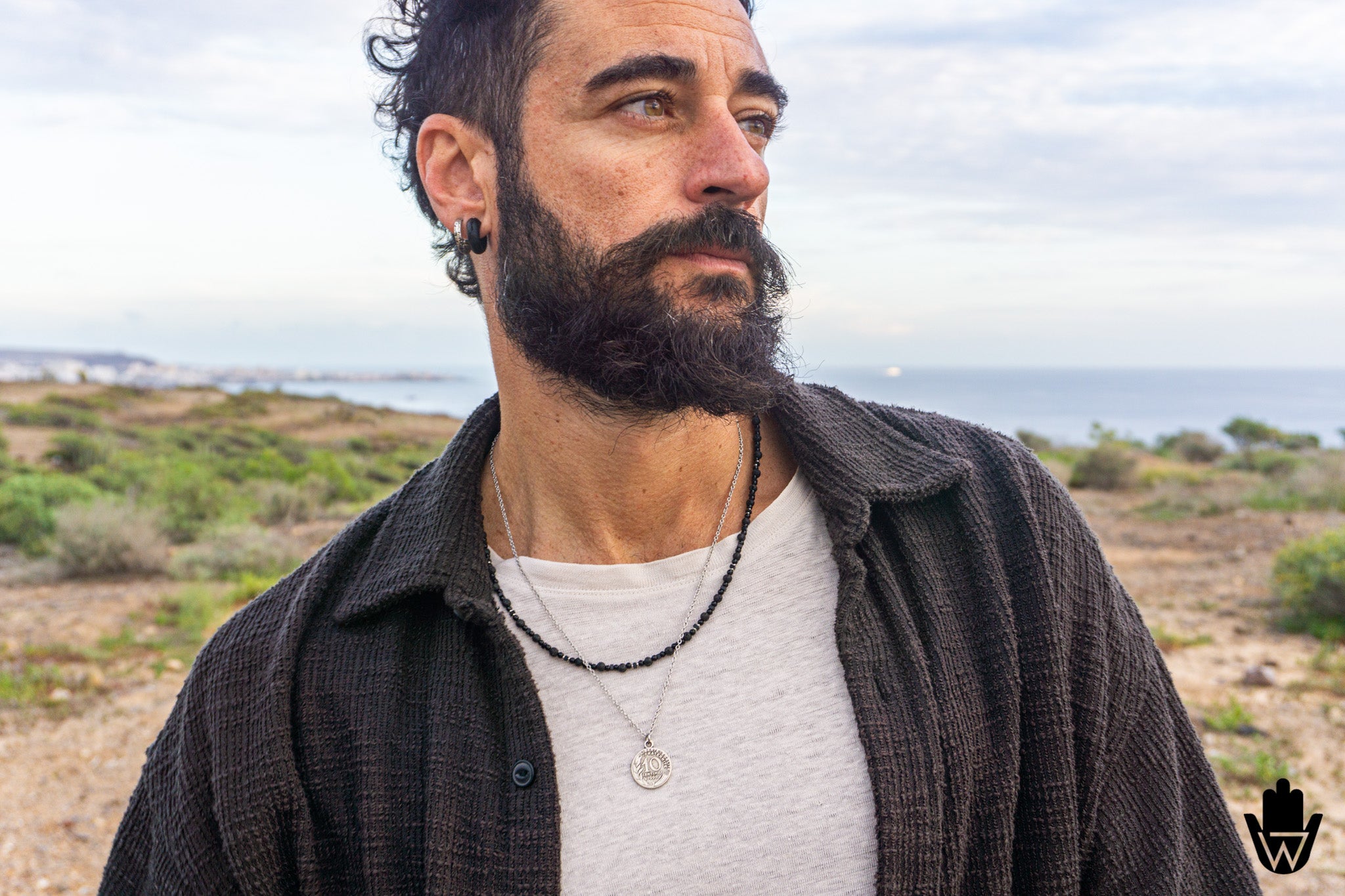 A man with a dark beard and curly hair wearing a dark textured button-up shirt over a light gray t-shirt, with layered necklaces, photographed outdoors against a blurred coastal landscape.
