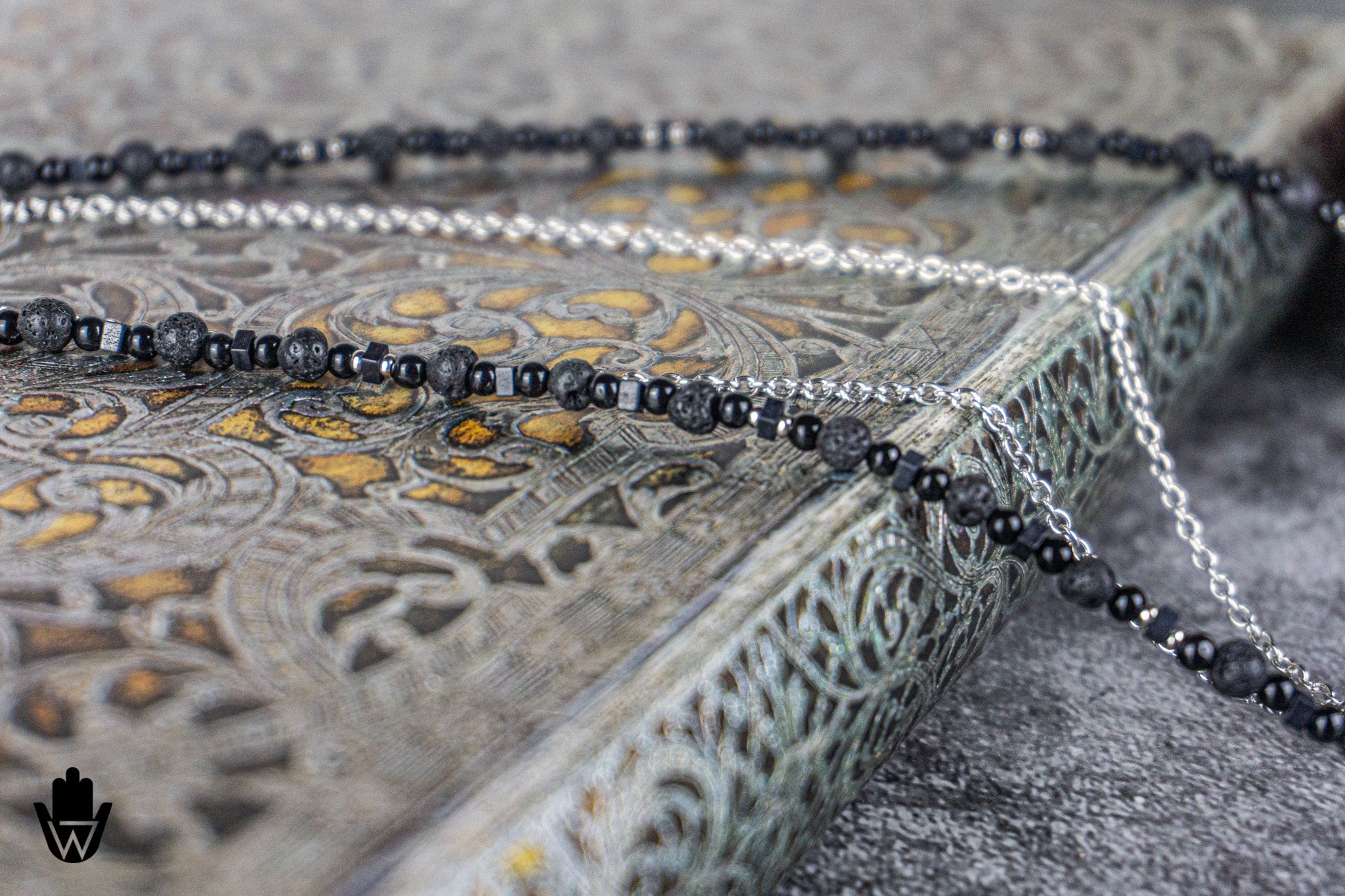 A close-up of a beaded necklace draped over an ornate engraved tray. The necklace features dark gray to black faceted round beads on one side and silver-toned round beads on the other, with a silver chain visible behind.