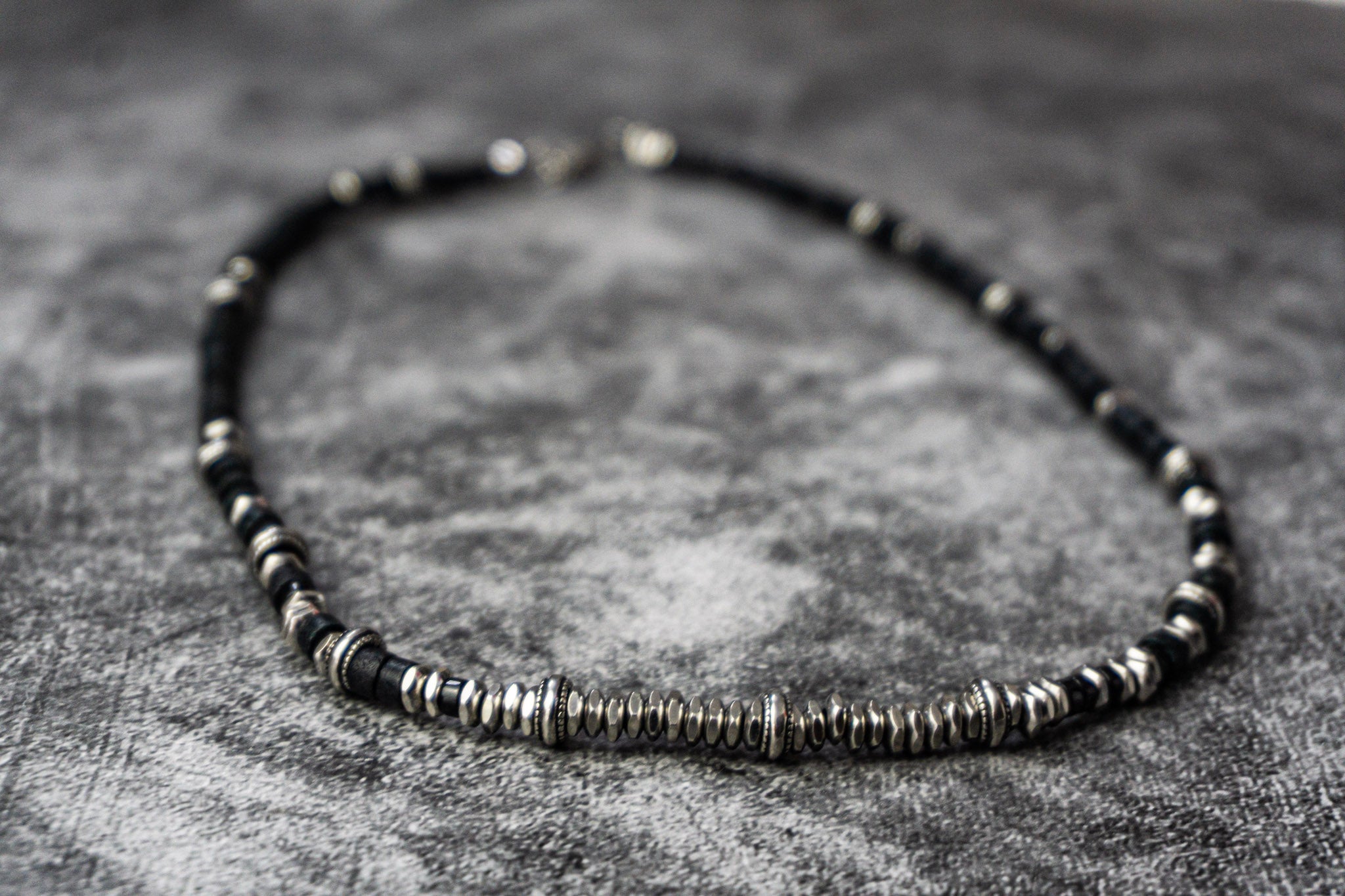 A beaded necklace featuring alternating black and silver metallic beads, arranged in a circle on a textured gray surface.