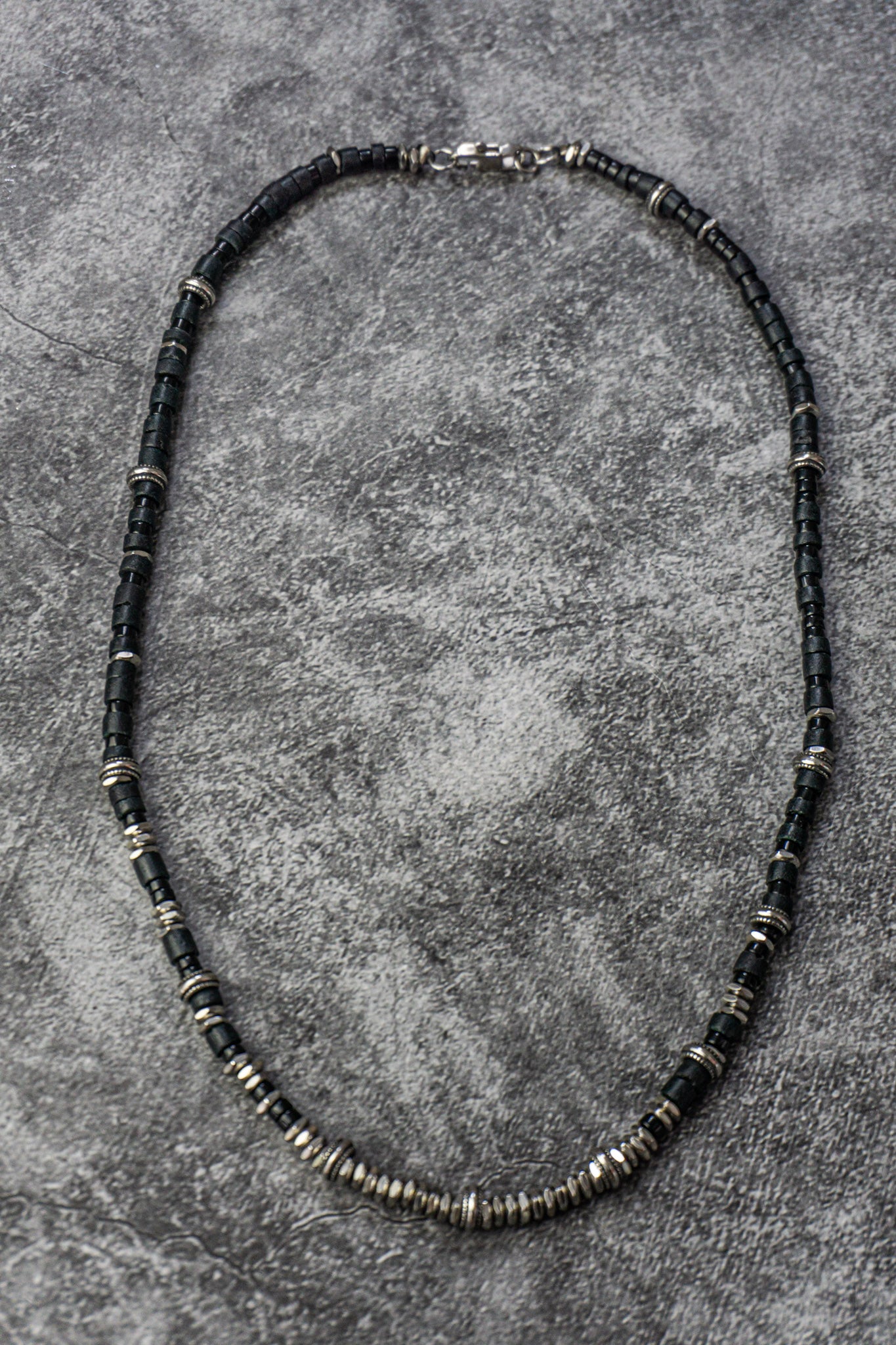 A beaded necklace made of small dark beads separated by metallic silver spacer rings, displayed on a textured gray surface.