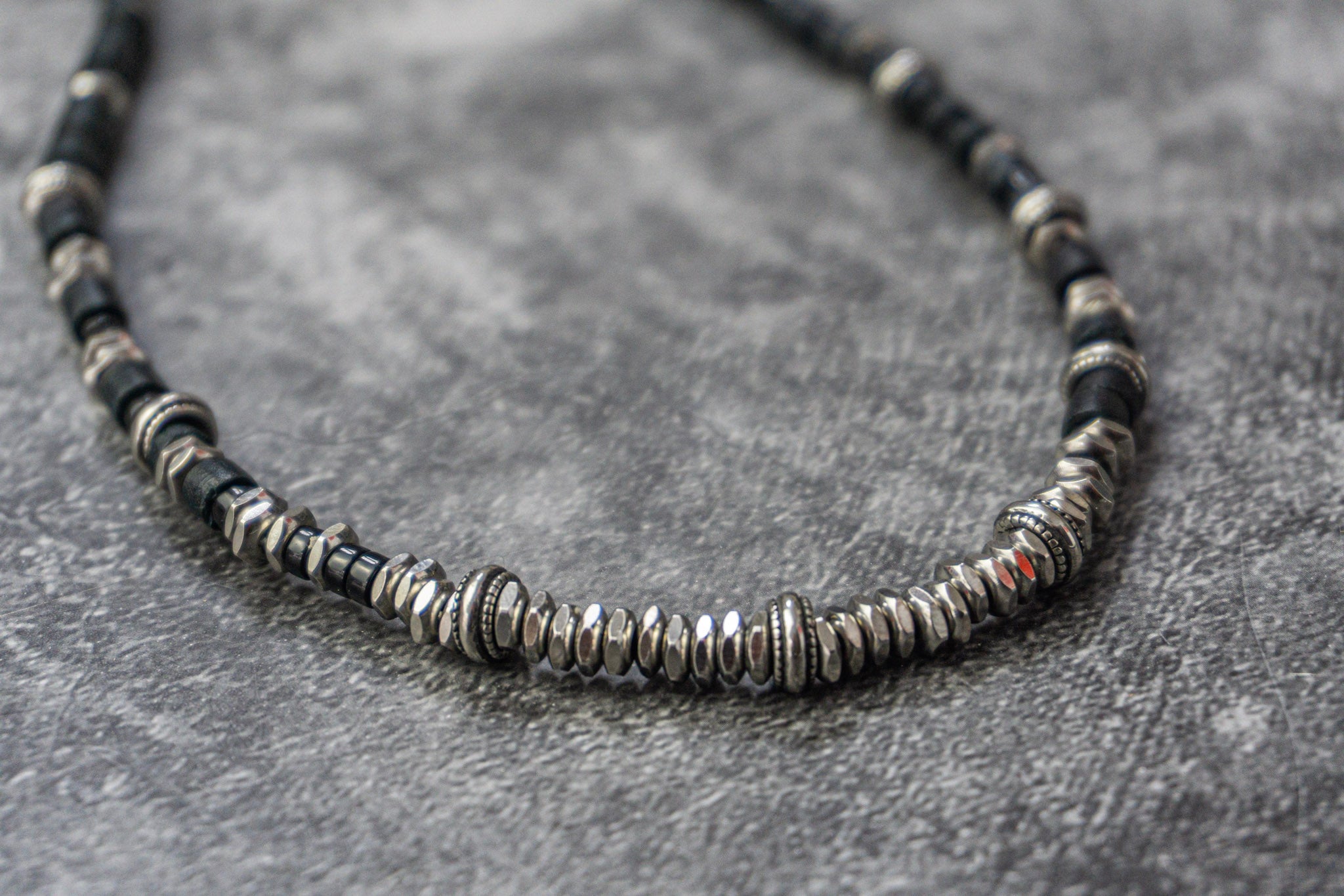A close-up of a beaded necklace with alternating black and silver-toned textured disc beads, resting on a textured gray surface.