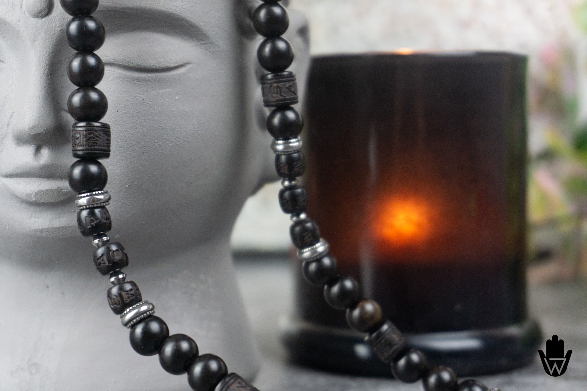 A close-up of a beaded necklace draped over a white Buddha head bust, with a dark amber glass candle holder and glowing orange candle in the softly blurred background.