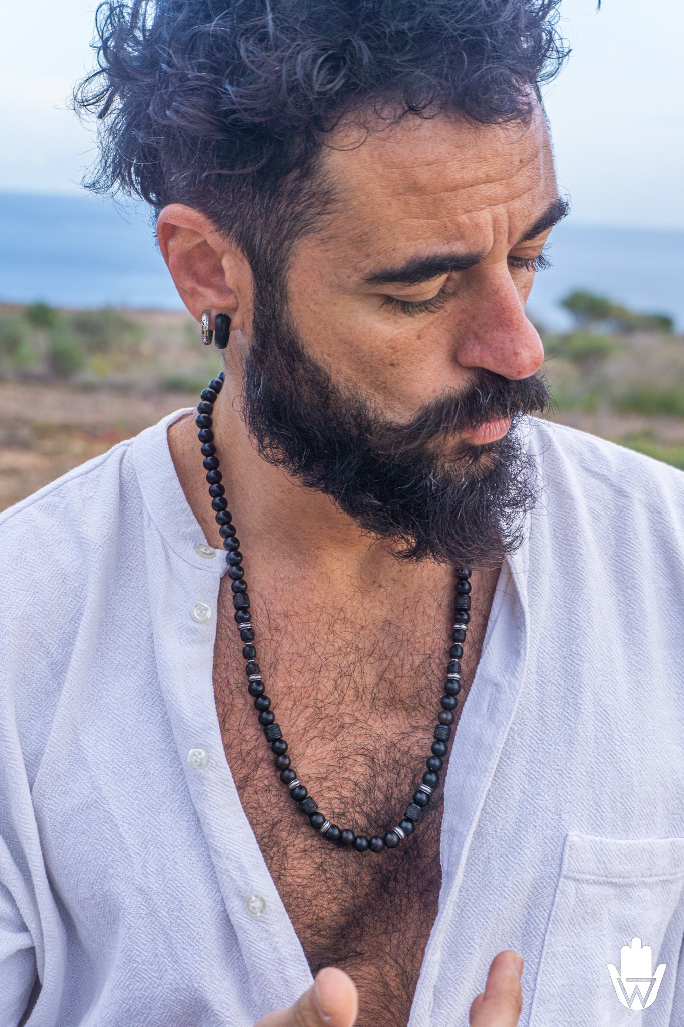 A close-up lifestyle shot of a man with a dark curly beard and mustache wearing a black beaded necklace and a black ear plug, wearing an open white shirt against an outdoor coastal background.