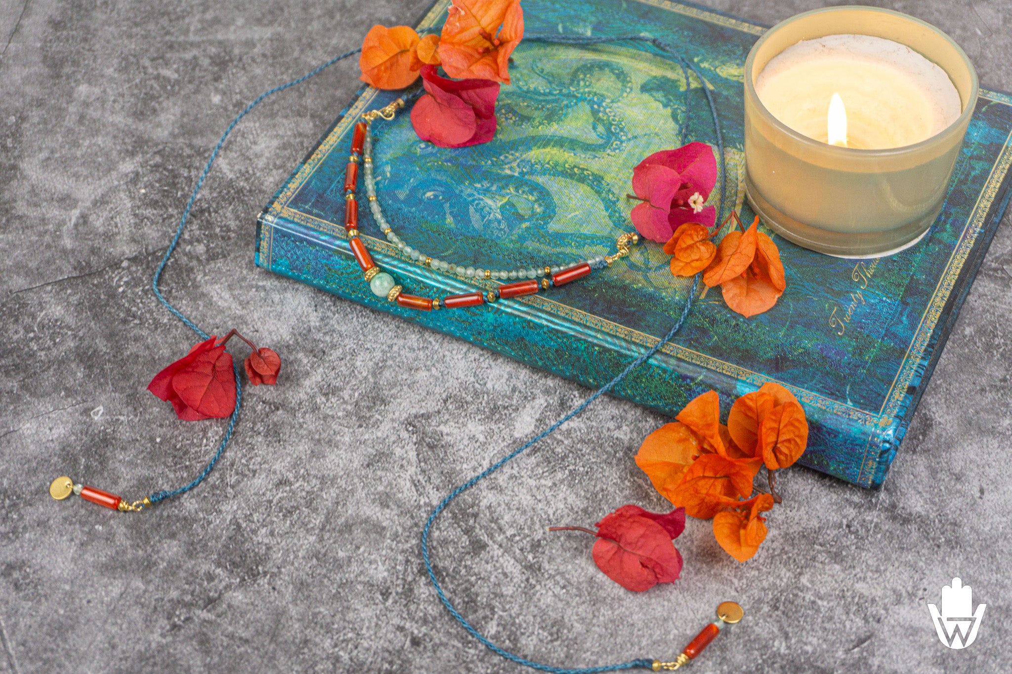 A beaded necklace with blue tail cords, red-orange beads, and small gold-toned accents is displayed on a textured gray surface next to a teal book, orange flowers, and a lit candle.