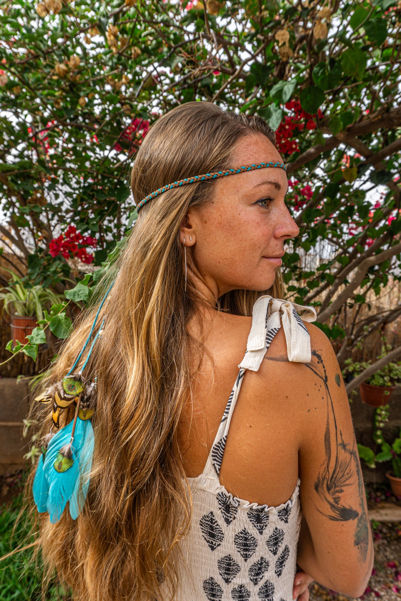 Woman with long hair and feathered headband standing outdoors with greenery in the background