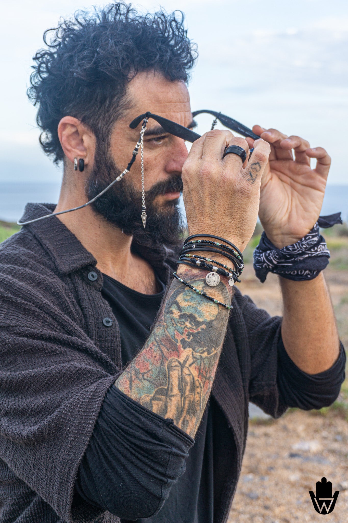 A man outdoors wearing black sunglasses with a silver chain detail, layered over a dark button-up shirt and black undershirt. He has a full beard, curly hair, and visible tattoos on his arm, with a bandana on his wrist.