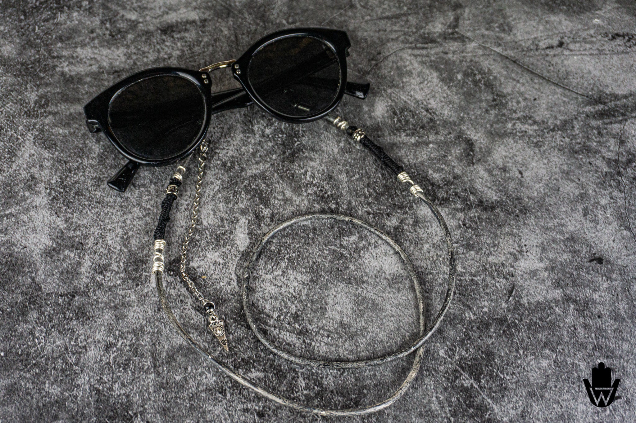 Black round sunglasses with a long silver chain strap featuring decorative metallic beads and charms, laid out on a textured gray surface.