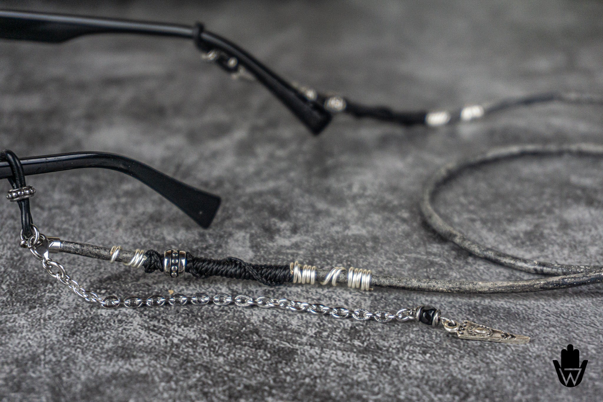 Black and silver metal chain glasses strap with decorative silver beads and a silver chain link extension, shown attached to black eyeglass arms on a textured gray background.