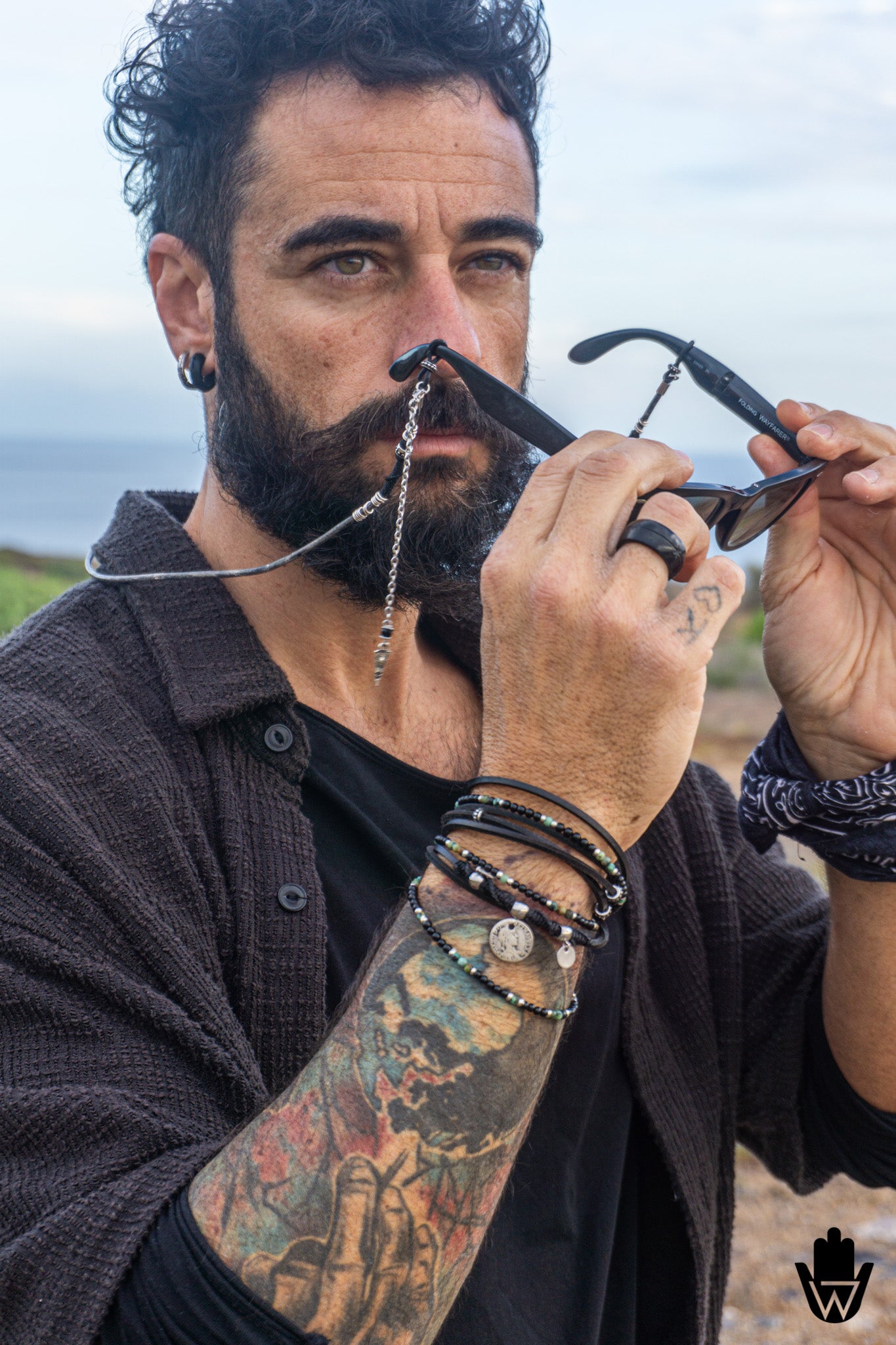 A close-up lifestyle shot of a bearded man with tattoos holding a pair of black sunglasses with a silver chain strap, wearing multiple leather bracelets on his wrist, against an outdoor beach background.