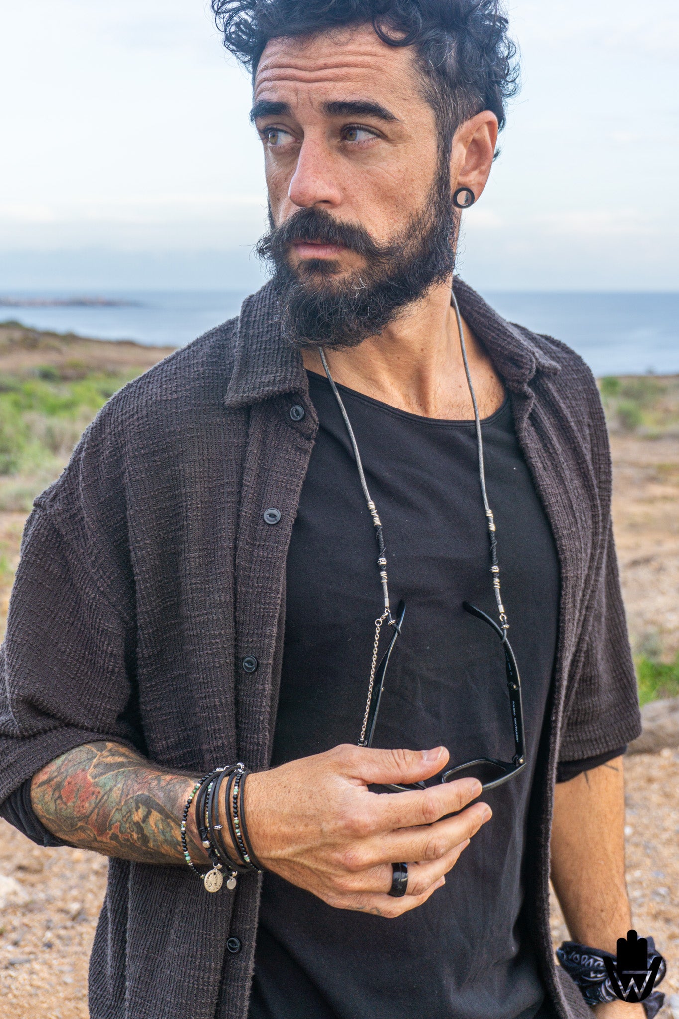 A man with a beard and tattoos wearing a dark brown textured button-up shirt over a black t-shirt, standing outdoors near the ocean with sunglasses hanging from a silver beaded necklace.