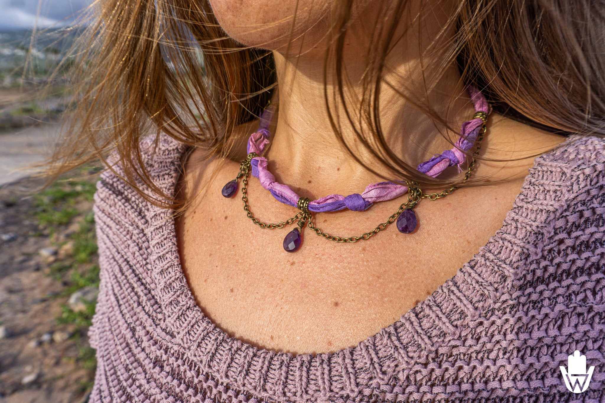 A close-up of a woman wearing a bohemian-style necklace with purple fabric beads, purple teardrop gemstones, and bronze chain links, set against an outdoor background.
