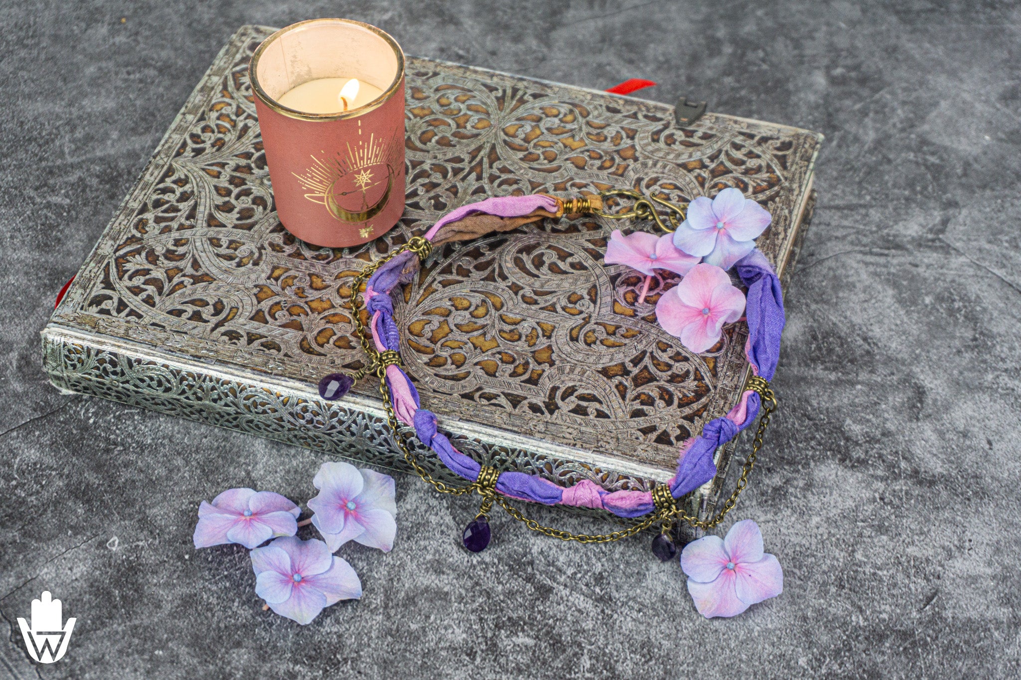 A bohemian-style beaded necklace with purple and pink fabric strands, dark purple beads, and gold-toned metal details, styled on a silver ornate tray next to a lit pink candle with scattered purple flower petals on a gray stone surface.