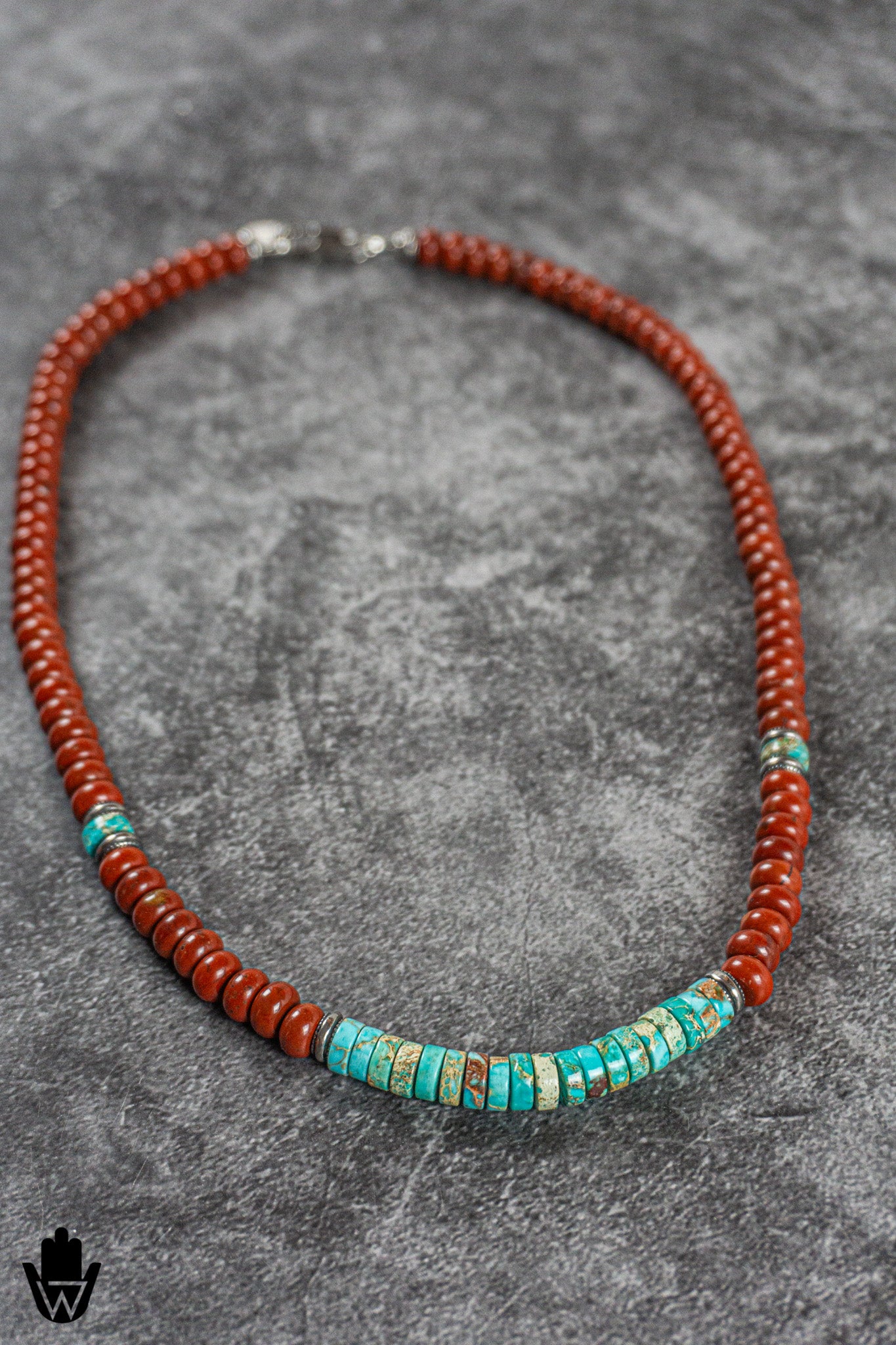 A beaded necklace with predominantly reddish-orange round beads and a central cluster of turquoise-blue rectangular beads, displayed on a textured gray background.