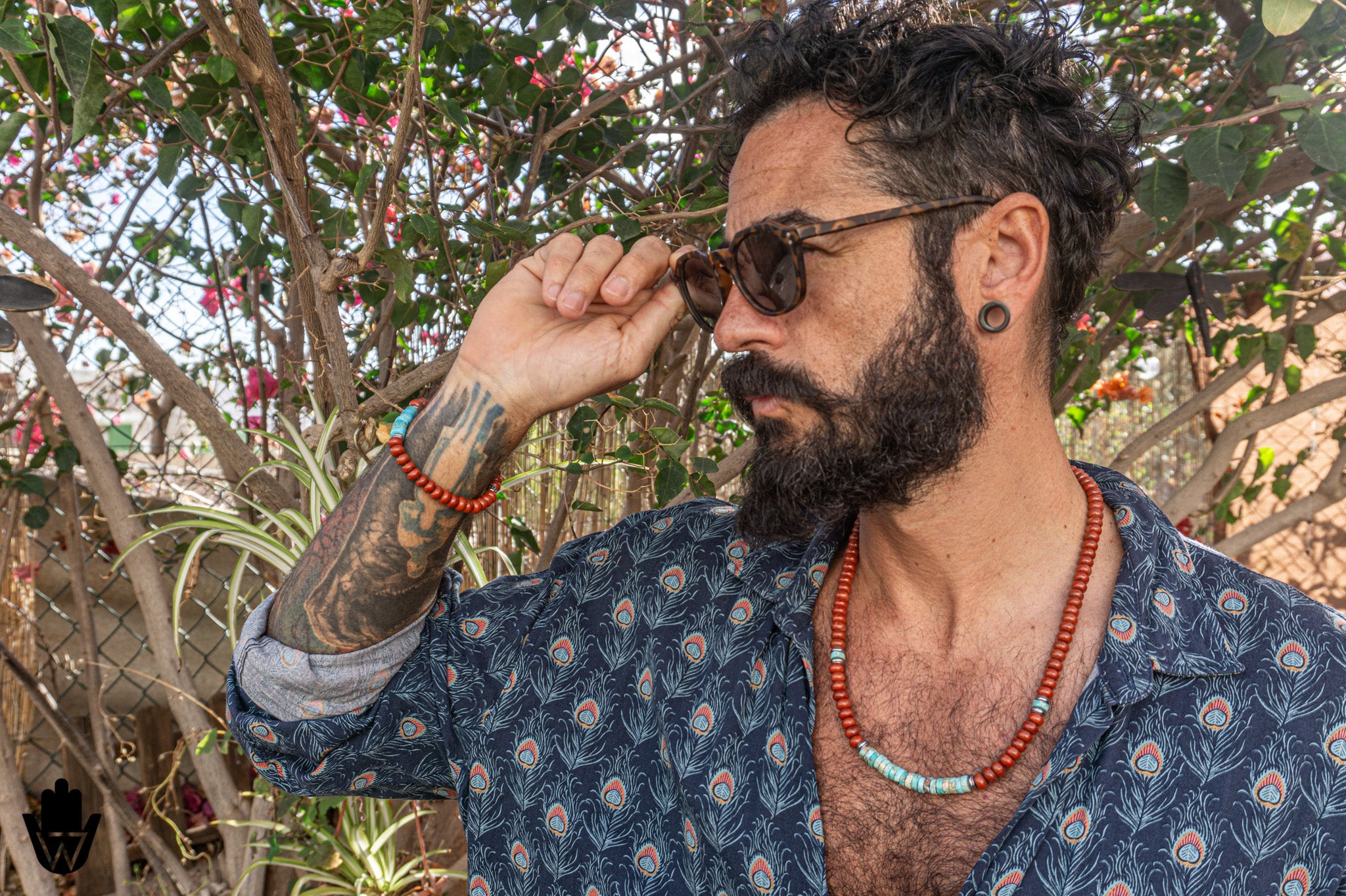 A man with a beard and tattoos wearing round sunglasses, a blue patterned shirt, and red beaded necklaces and bracelets, standing outdoors in front of leafy trees.