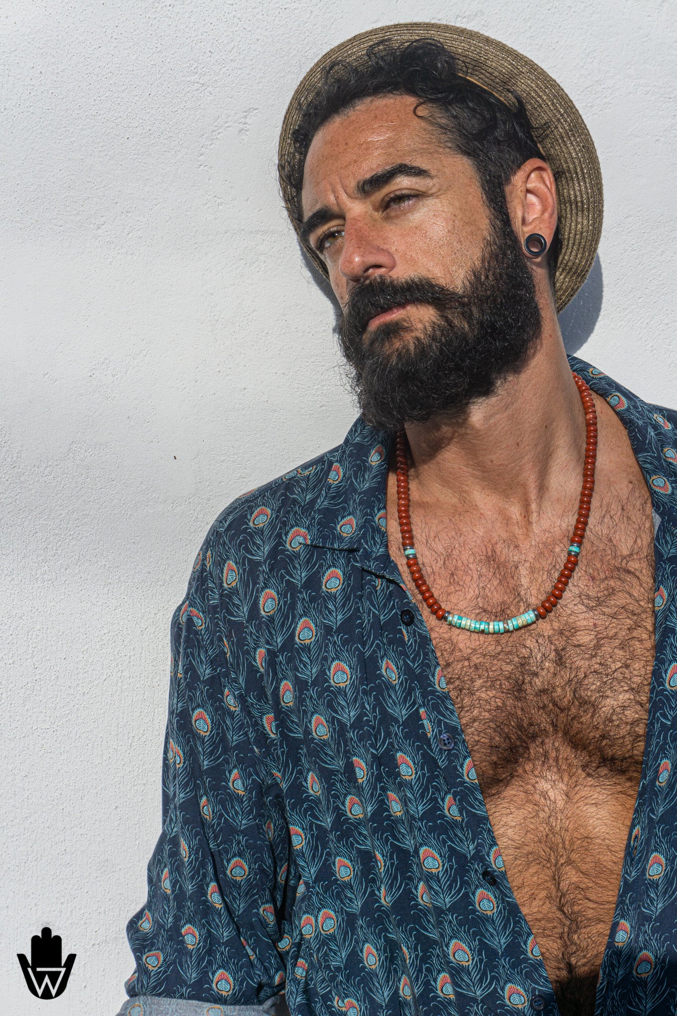 A man with a dark beard and mustache wearing a straw hat, a blue patterned button-down shirt, and a red beaded necklace, posing against a white wall.