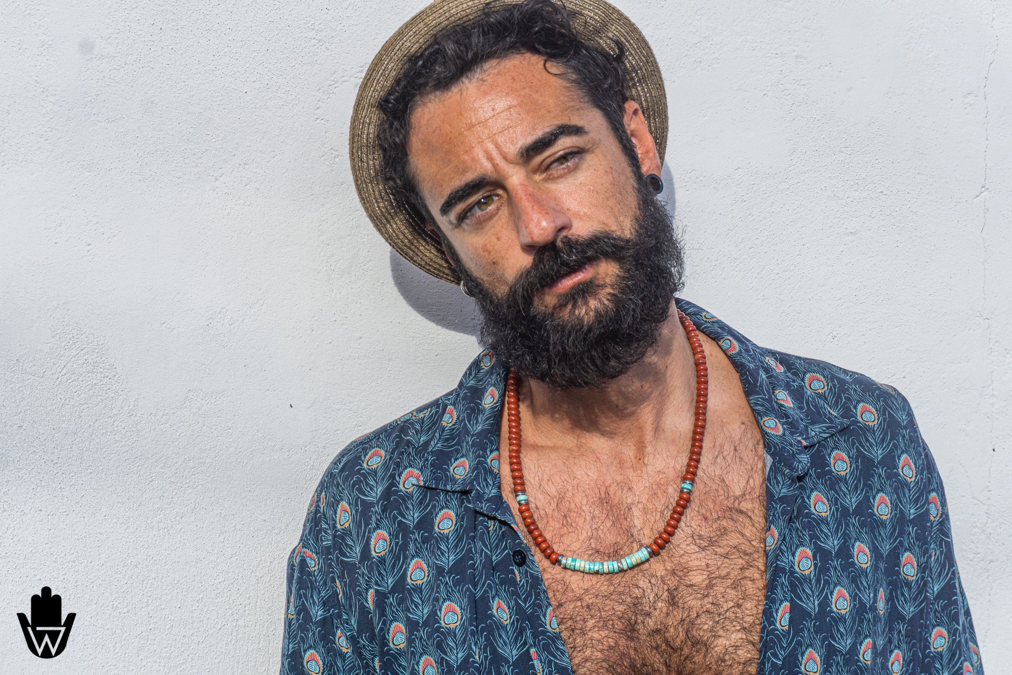 A man with a dark beard and mustache wearing a straw hat, a blue patterned button-down shirt, and a red beaded necklace, posing against a white textured wall.