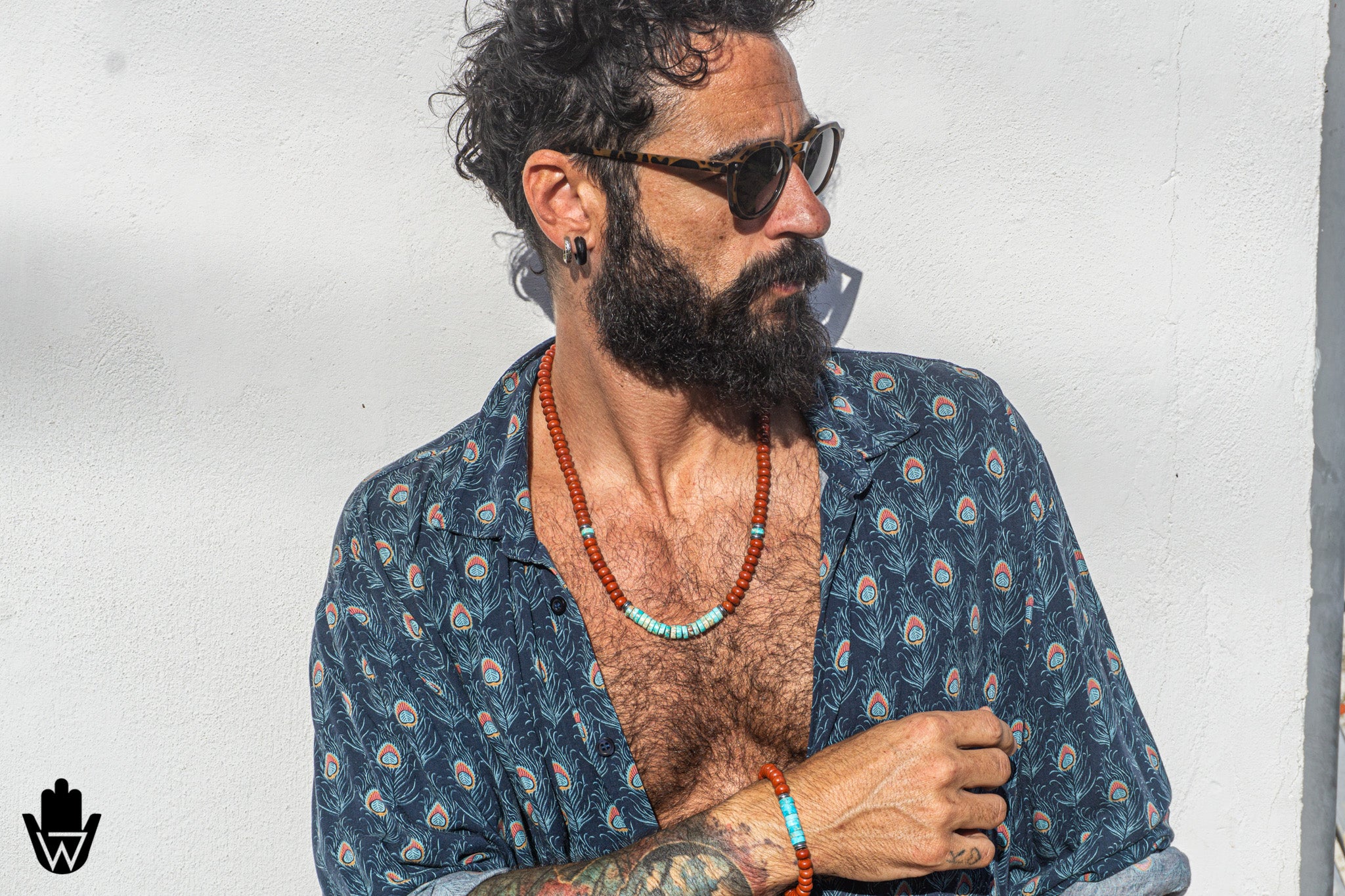 A man with a dark beard and sunglasses wearing an open navy blue patterned shirt, accessorized with a turquoise and red beaded necklace and matching bracelet, posing against a white wall.