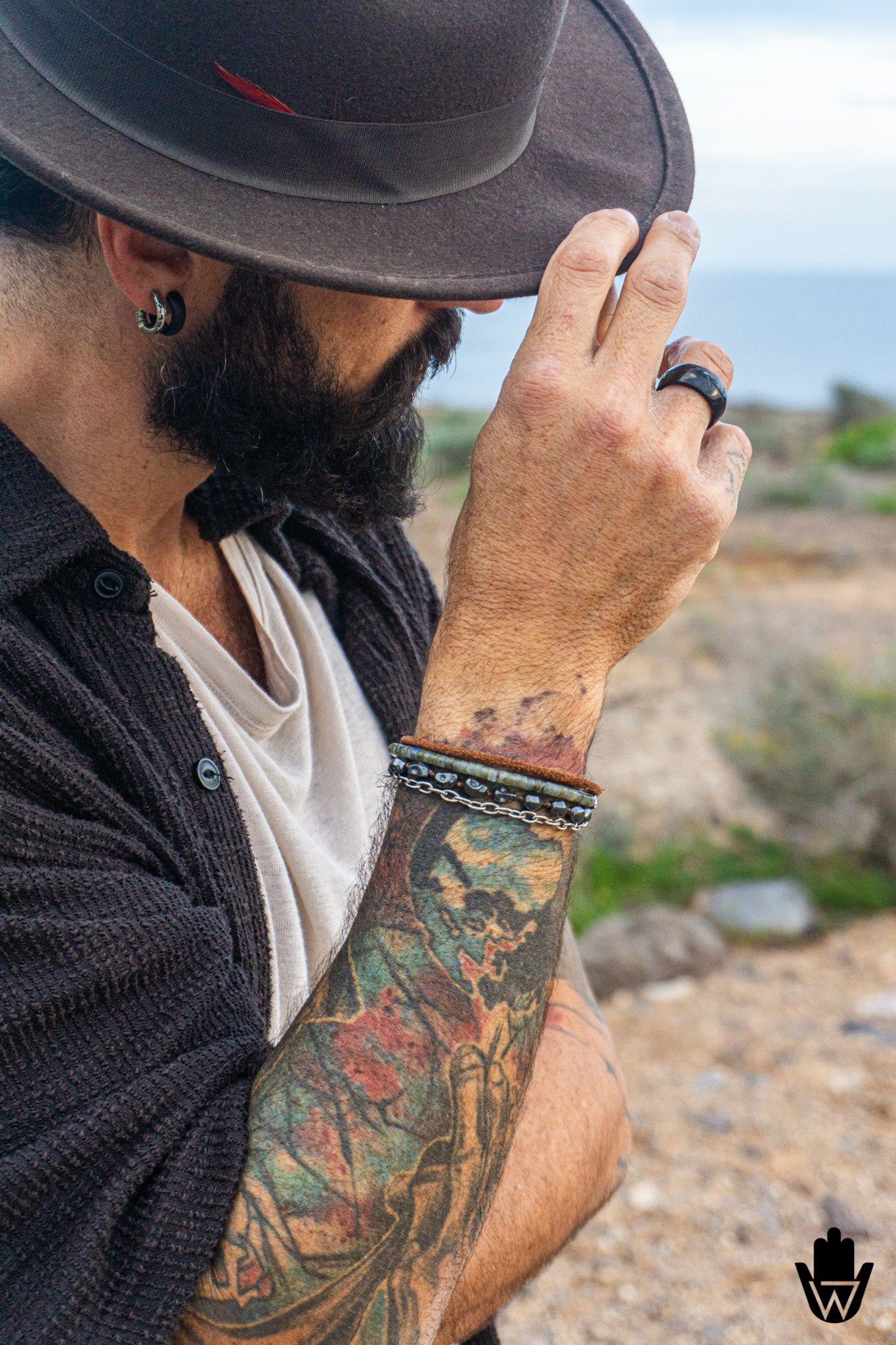 A close-up lifestyle shot of a bearded man wearing a black wide-brim hat, black shirt, and a colorful tattooed arm, showing a black leather ring on his finger and a beaded bracelet set on his wrist, with a blurred outdoor background.