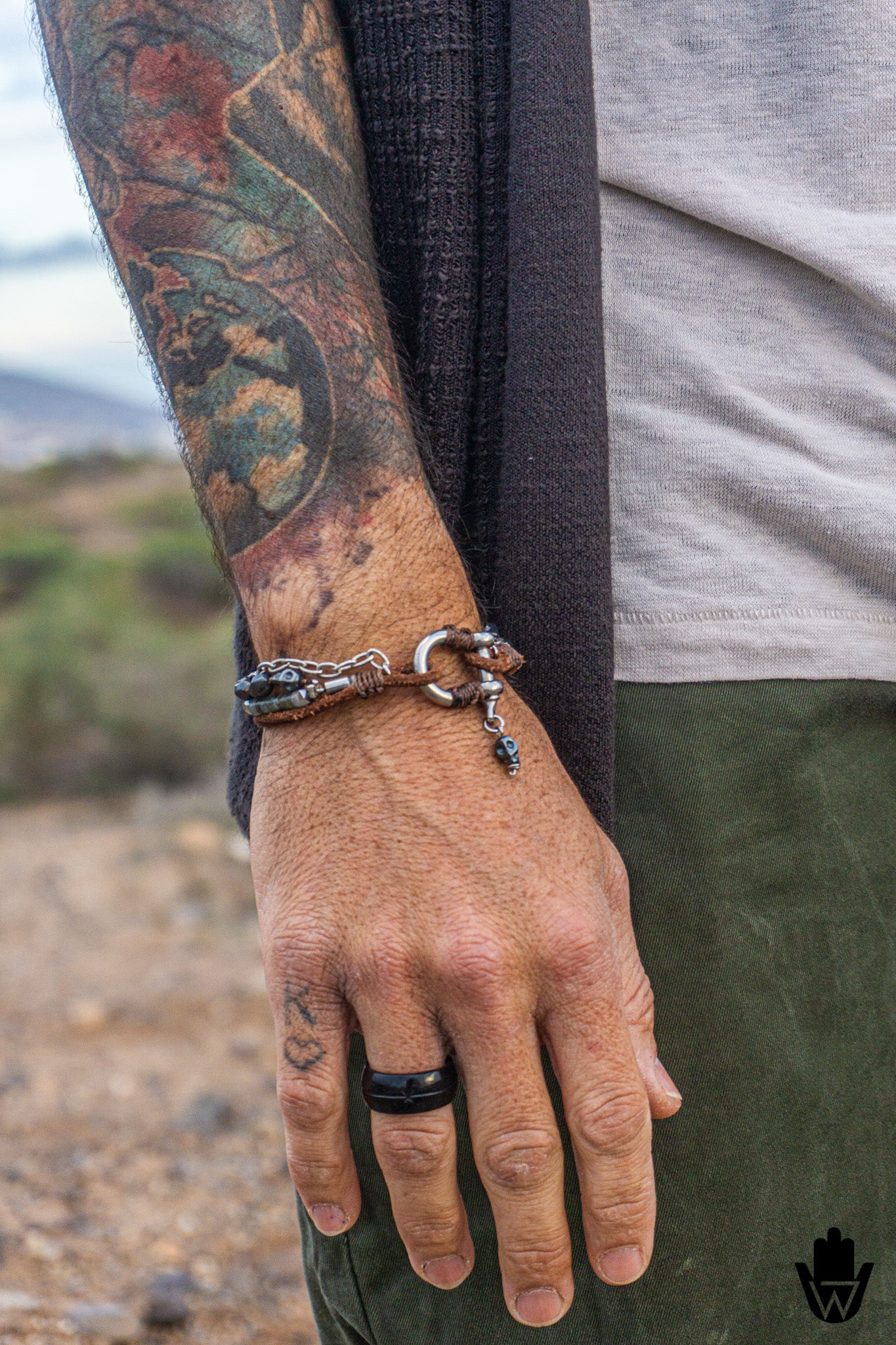 A close-up of a person's tattooed forearm and hand wearing a silver-toned metal bracelet with a circular ring detail and small dangling charm, along with a black ring on the finger.