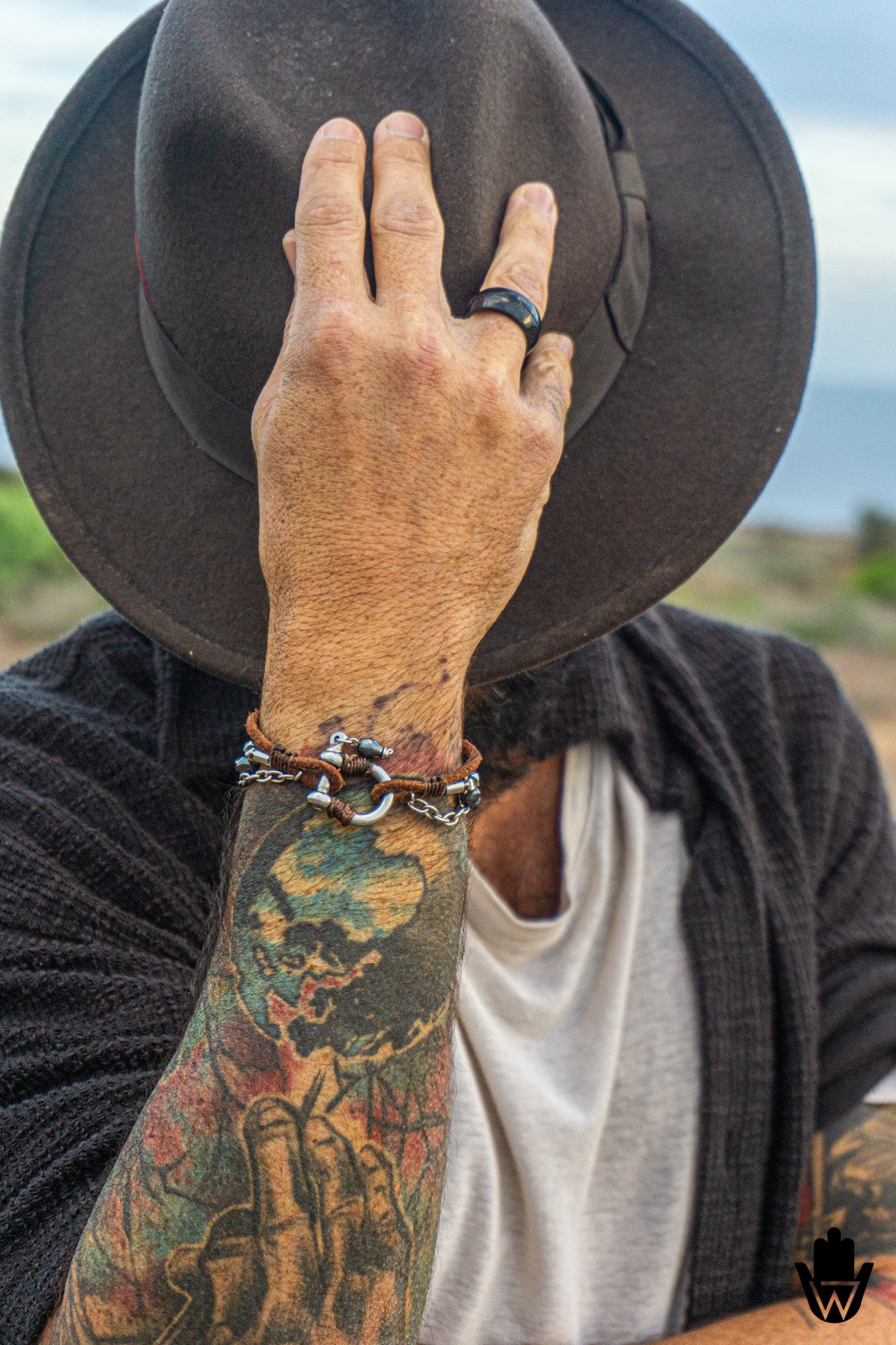 A close-up lifestyle shot of a tattooed man wearing a dark brown wide-brim hat, a black knit cardigan over a white t-shirt, and a silver chain bracelet with leather on his wrist.