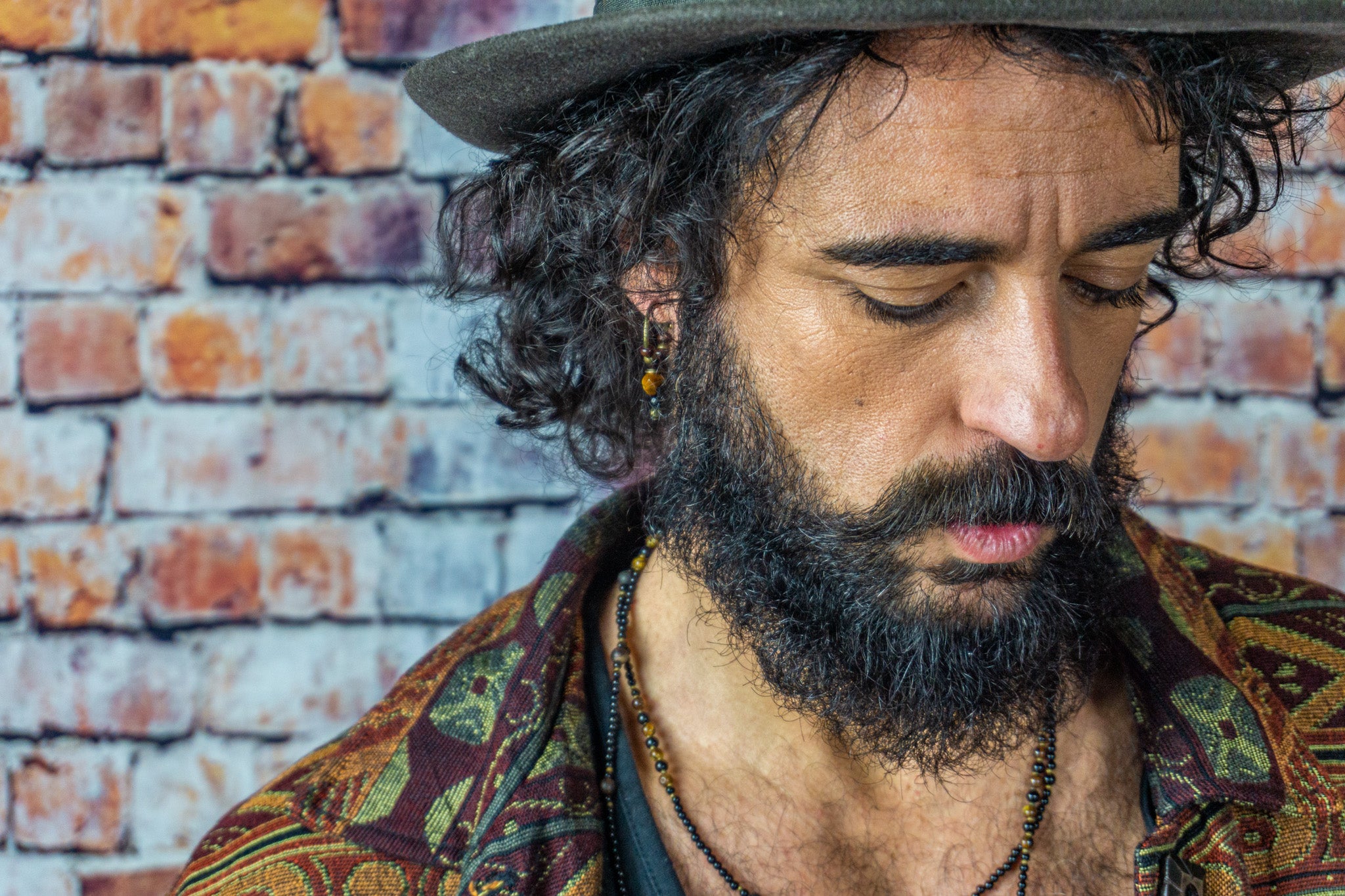 A long haired man with a hat, colorful vest, long layered onyx necklace and a tiger eye earring.
