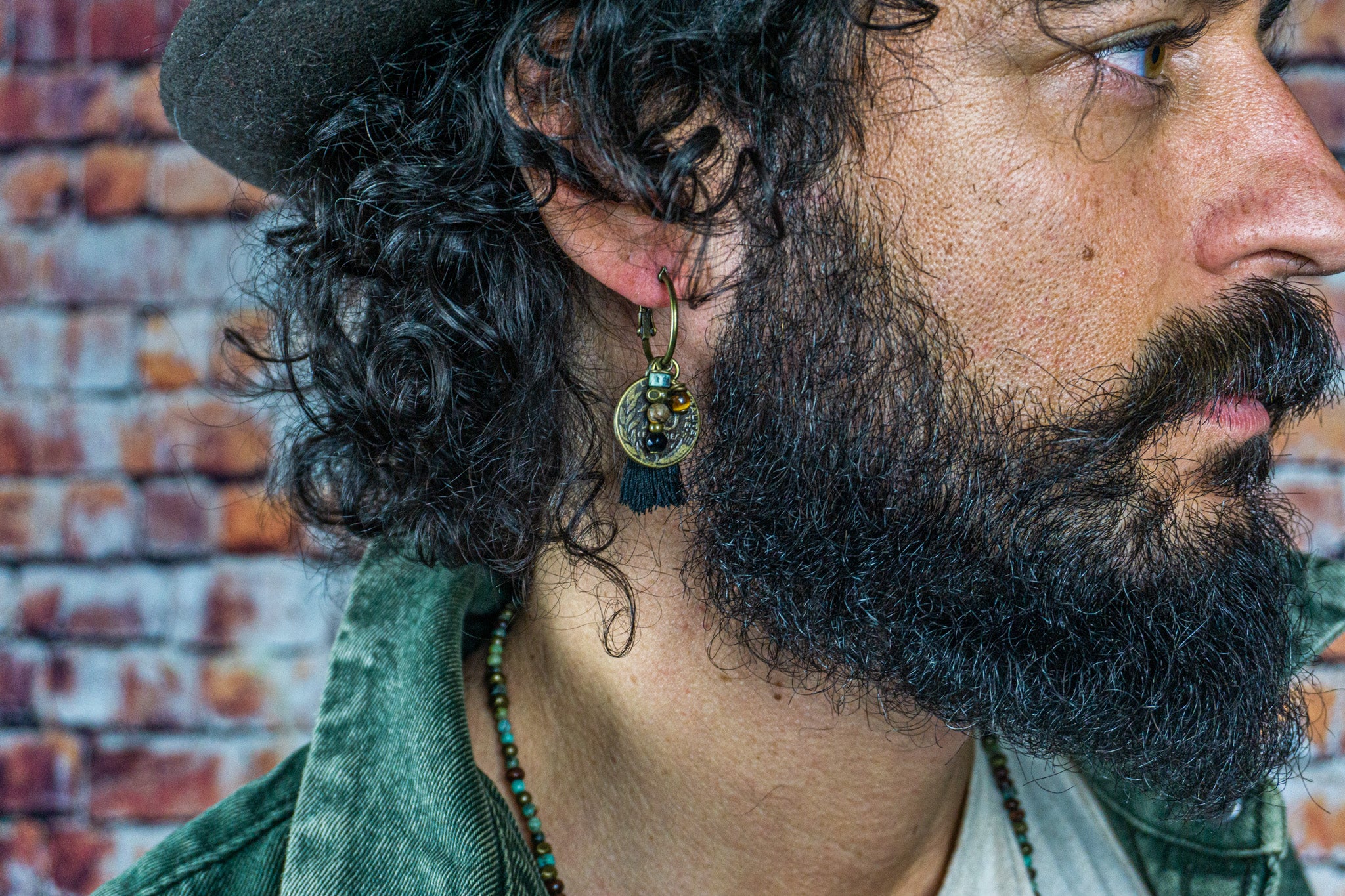A long haired man with a beard and a bronze hoop earring with an ancient coin, a black tassel and a strand of gemstones. wander jewellery 