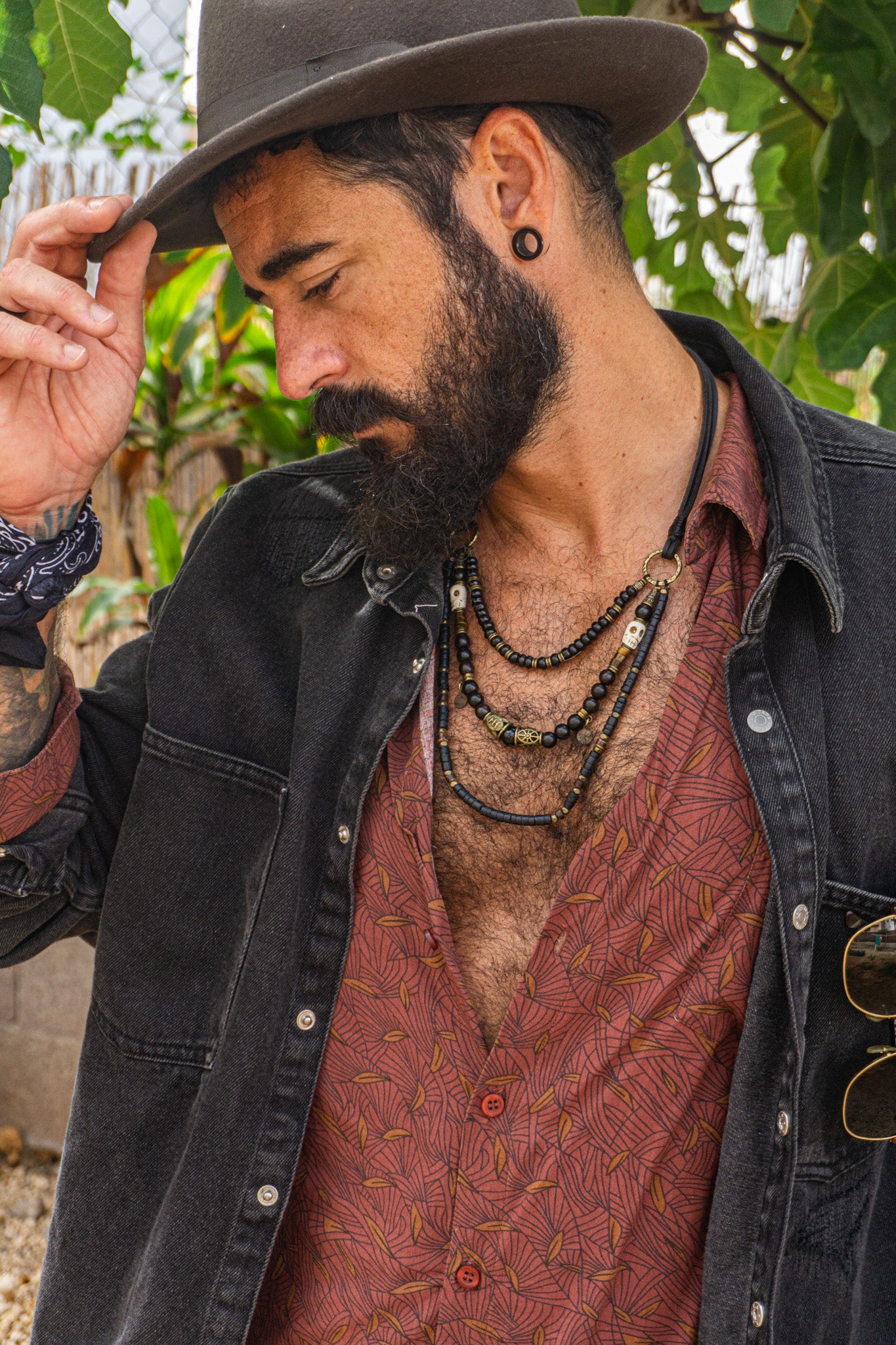 Man wearing a hat, black jacket, and patterned shirt with a layered black necklace