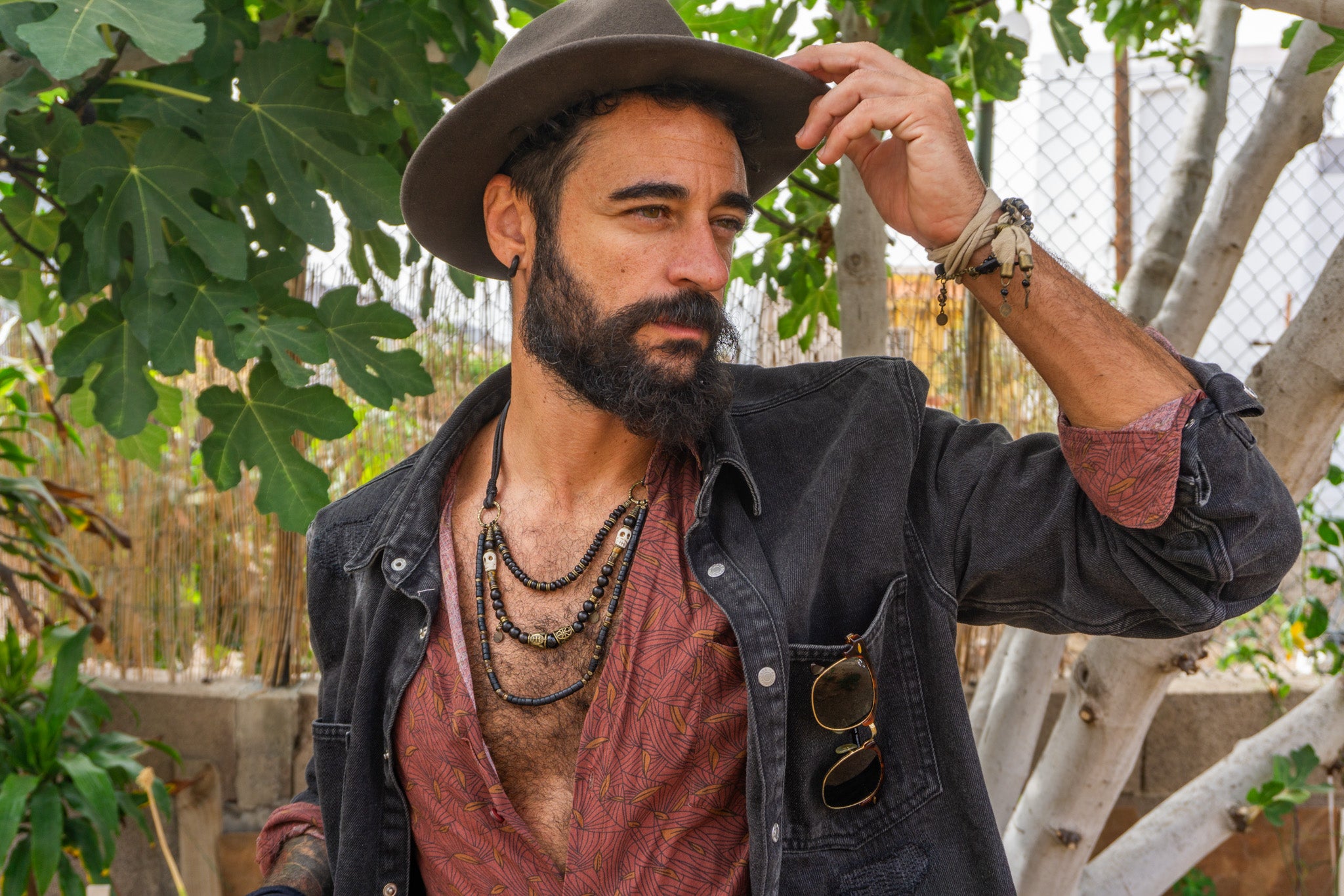 Man with a beard wearing a hat and layered necklaces outdoors