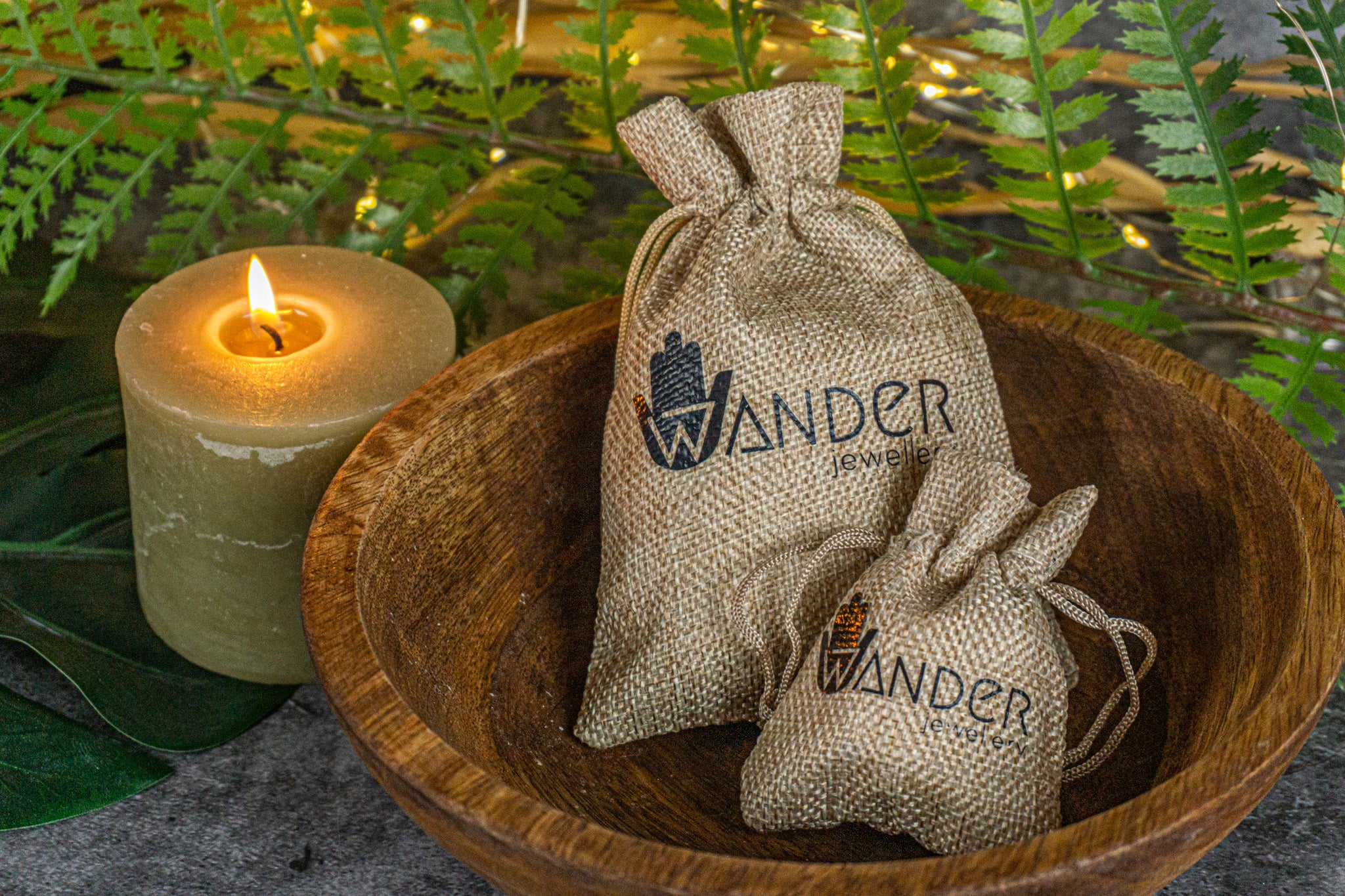 Two small burlap bags with 'Wanderjewellery' branding in a wooden bowl with a candle and greenery in the background.