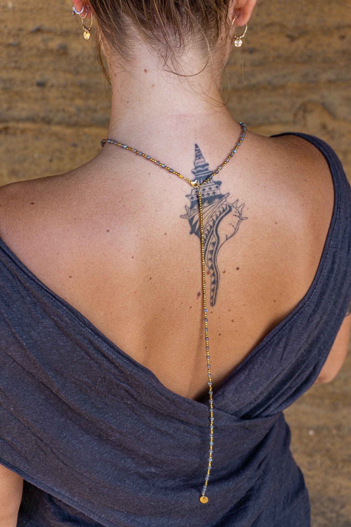 Women wearing a long drop back necklace with a detailed pendant, showing a tattoo on their back. Wander jewellery