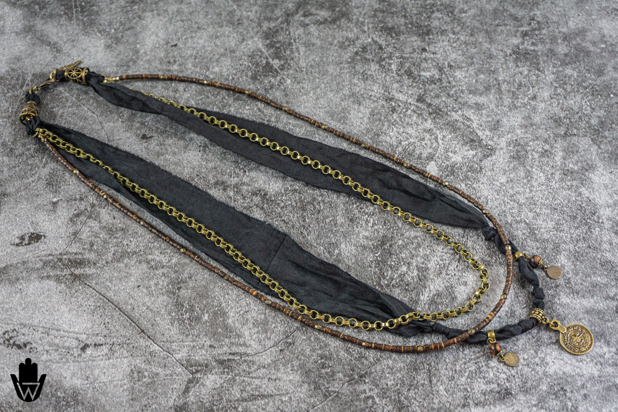A long layered necklace laid flat on a textured gray surface, featuring a black fabric ribbon, a gold-tone chain, and a bronze-tone beaded strand with a round coin charm.