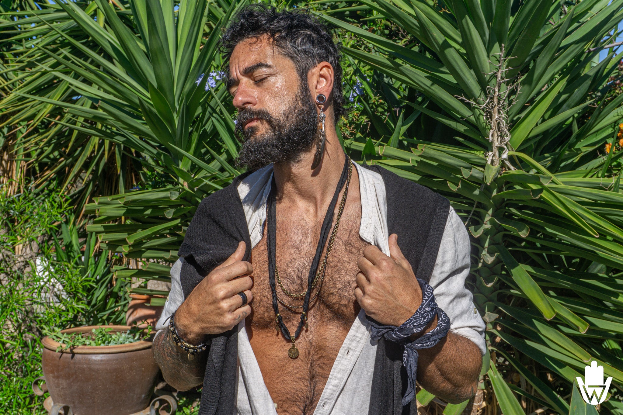 A bearded man with a black beard and ear piercings stands outdoors in front of green plants, wearing a white shirt with a black vest draped off his shoulders, layered with multiple black necklaces and bracelets.