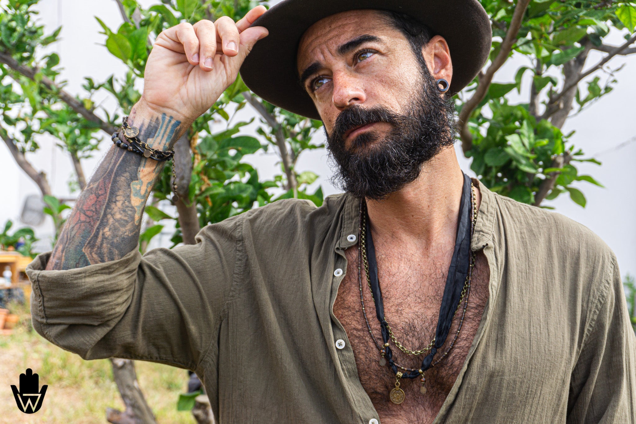A man with a full beard and tattoos wearing a dark brown wide-brim hat, an olive green open button-up shirt, and layered necklaces, posing outdoors in front of green trees.