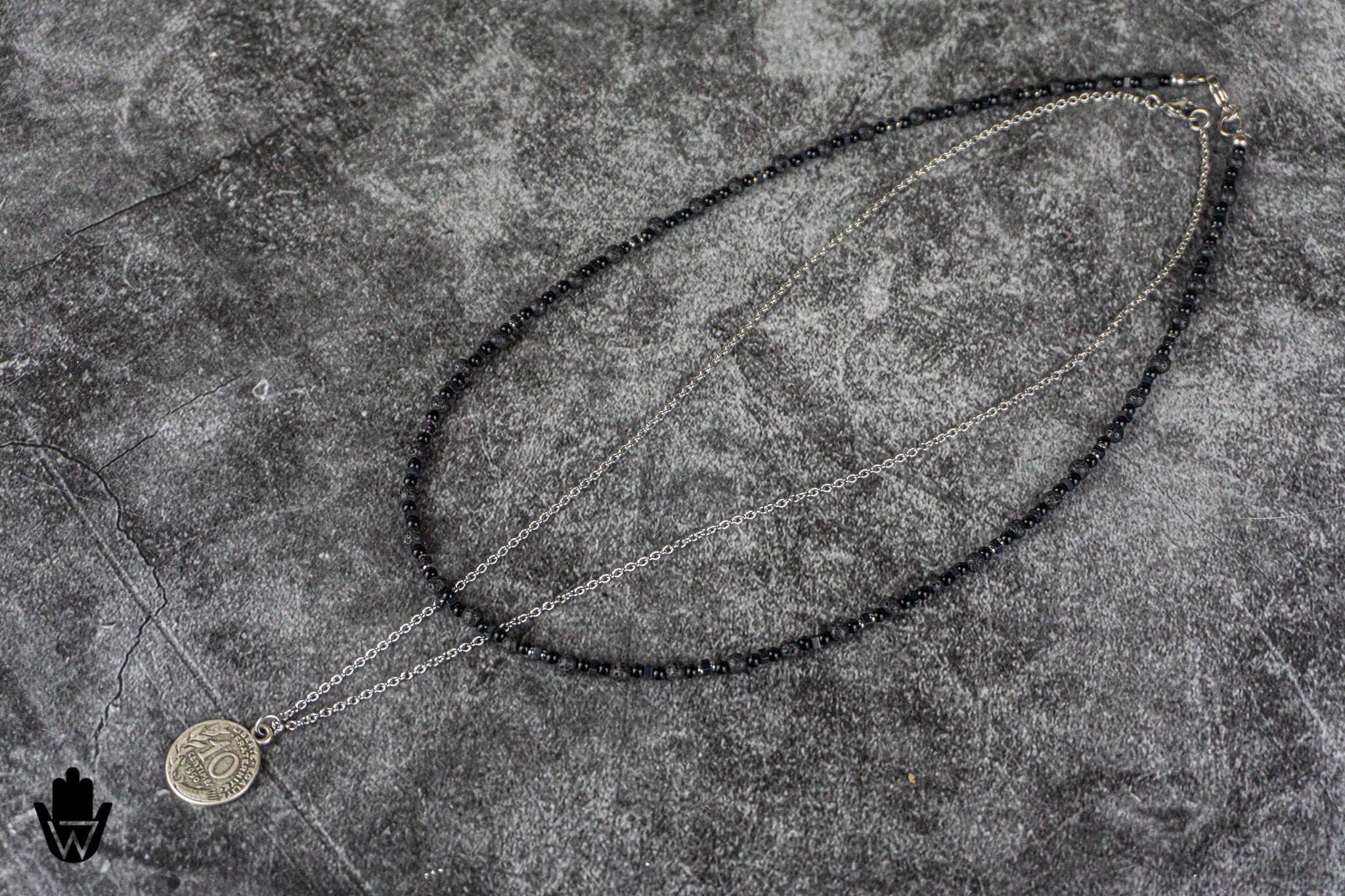 A double-strand necklace featuring a dark, beaded upper section and a silver-toned chain with a circular pendant, laid out on a textured gray stone surface.