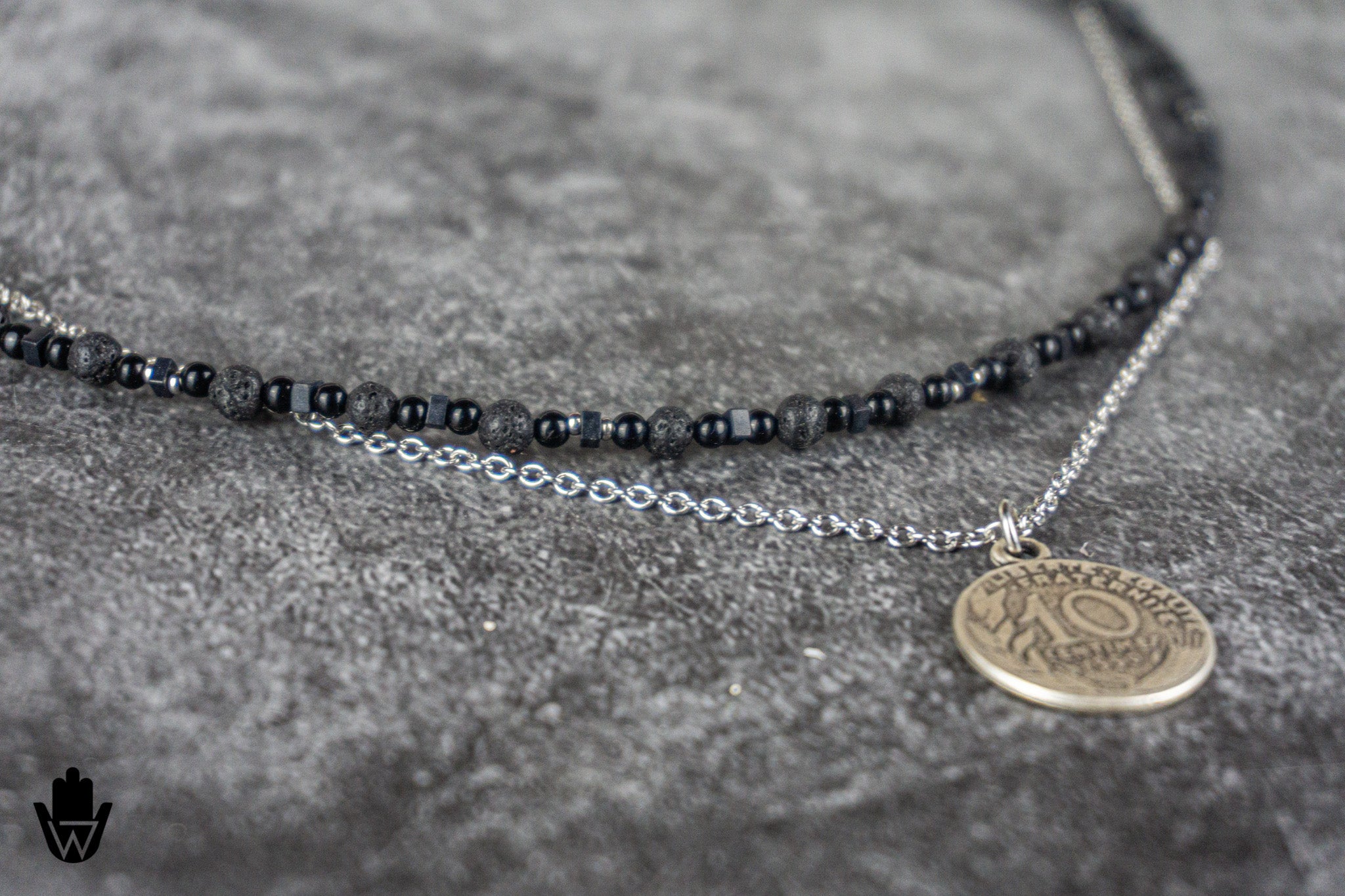 A close-up of a double-strand necklace featuring a beaded black chain and a silver chain with a round bronze-toned coin pendant, displayed on a textured gray surface.