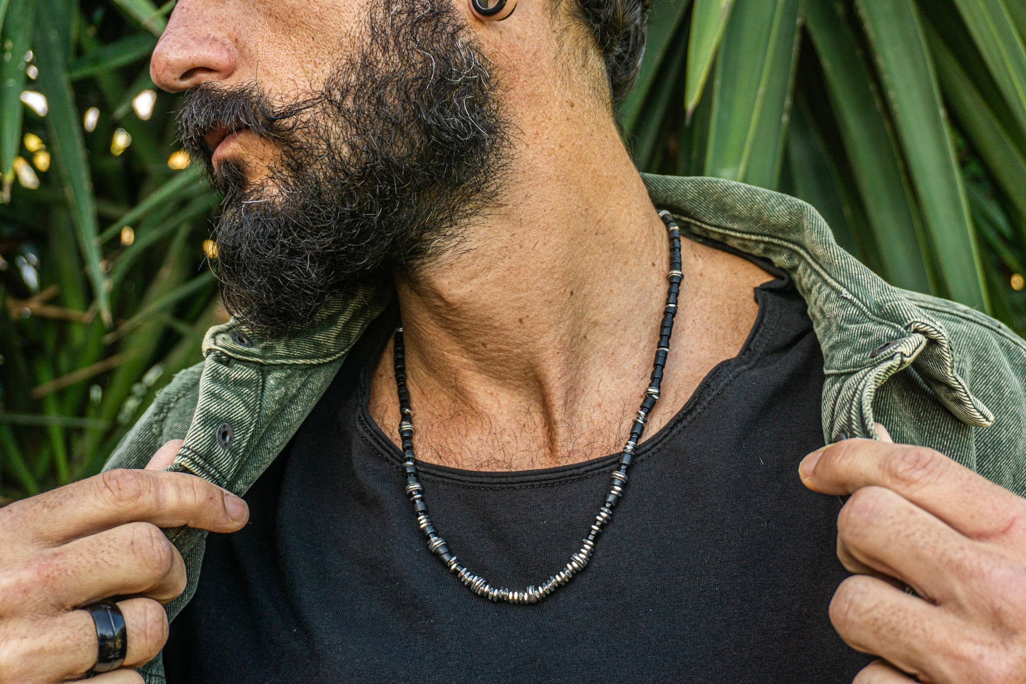 A close-up lifestyle shot of a man with a beard wearing a beaded necklace. The necklace features dark gray and metallic silver beads.
