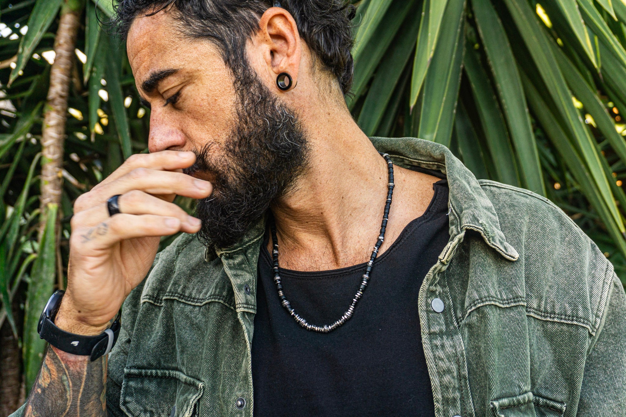 A man with a beard and black ear plugs wearing a black beaded necklace, black ring, and black watch, wearing an olive green denim shirt over a black t-shirt, with a background of green palm leaves.