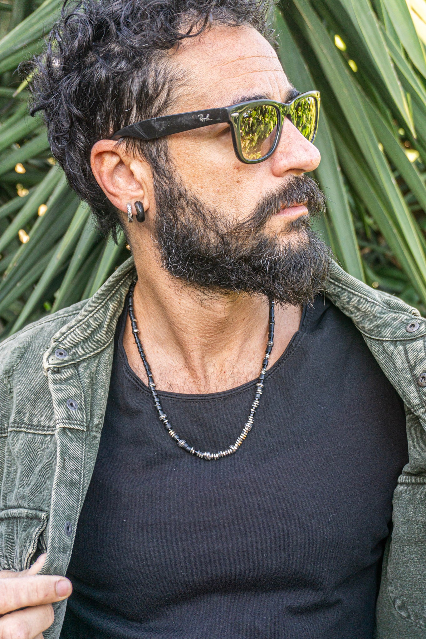 A man with a beard and curly hair wearing black sunglasses with gold mirrored lenses, a black t-shirt, and an open green denim jacket, set against a background of green palm leaves.
