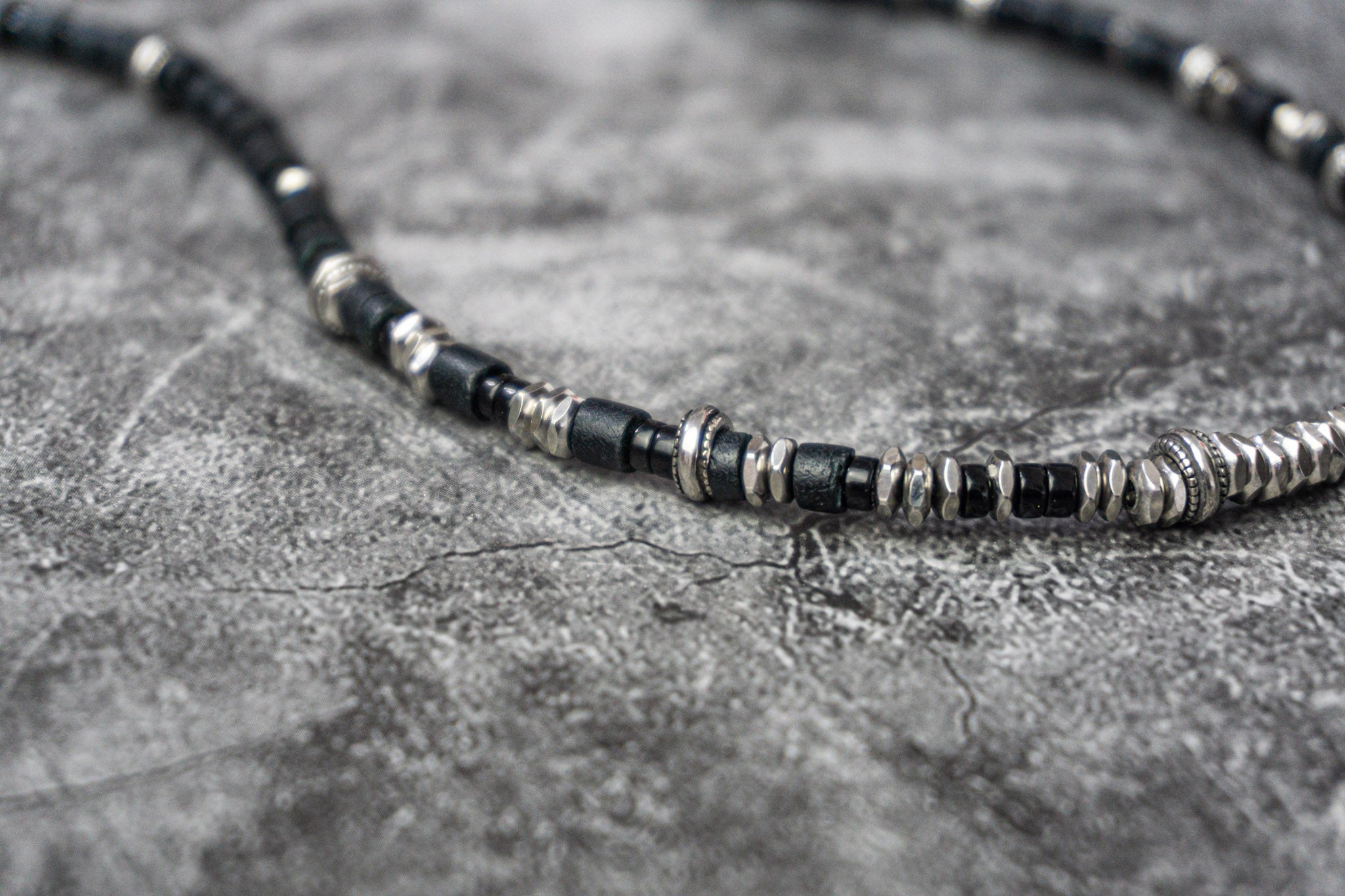 A close-up of a beaded necklace featuring alternating black and silver metallic beads, displayed on a textured gray stone surface.