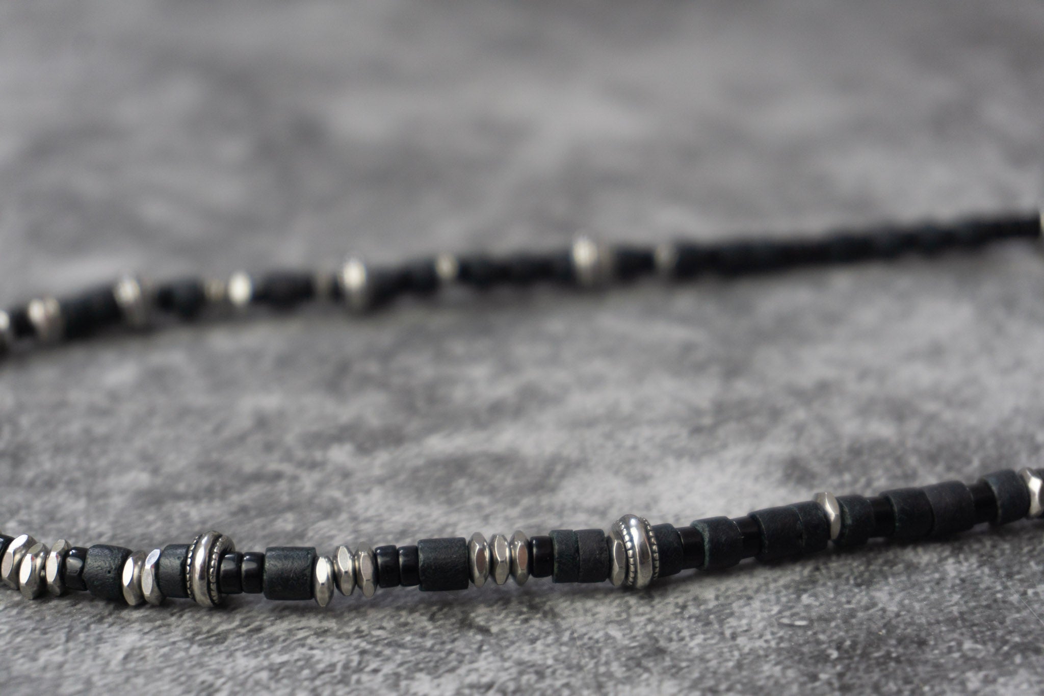 A close-up of a beaded necklace featuring alternating matte black and polished silver cylindrical beads, resting on a textured gray surface.