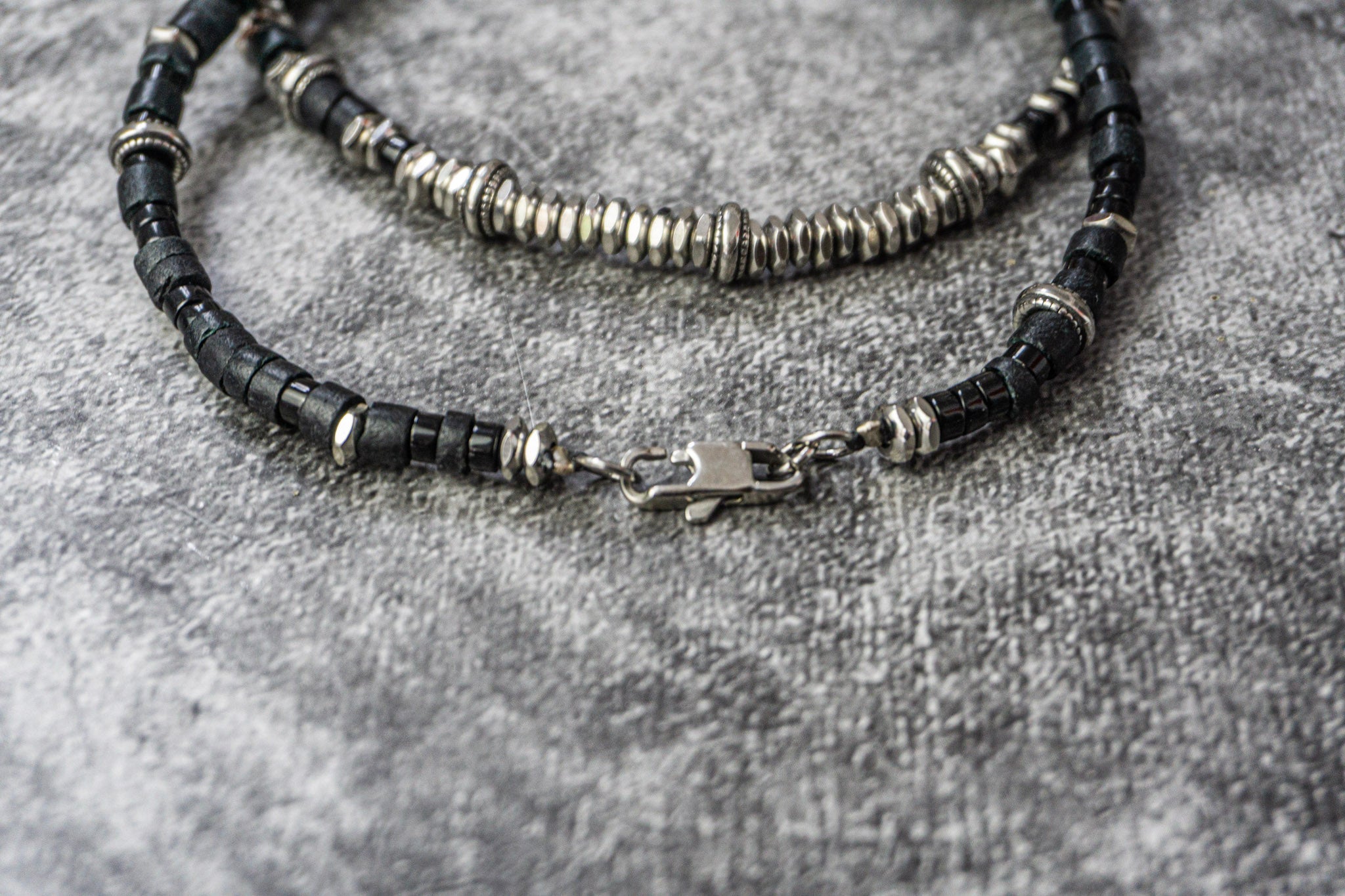 A close-up of a beaded bracelet or necklace with black faceted beads and silver-toned spacer rings and clasp, laid on a textured gray surface.