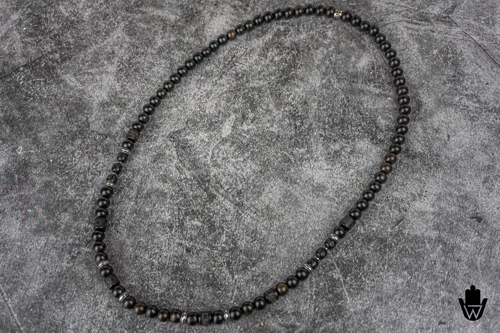 A beaded necklace made of small dark round beads with subtle metallic sheen, arranged in an oval shape on a textured gray surface.