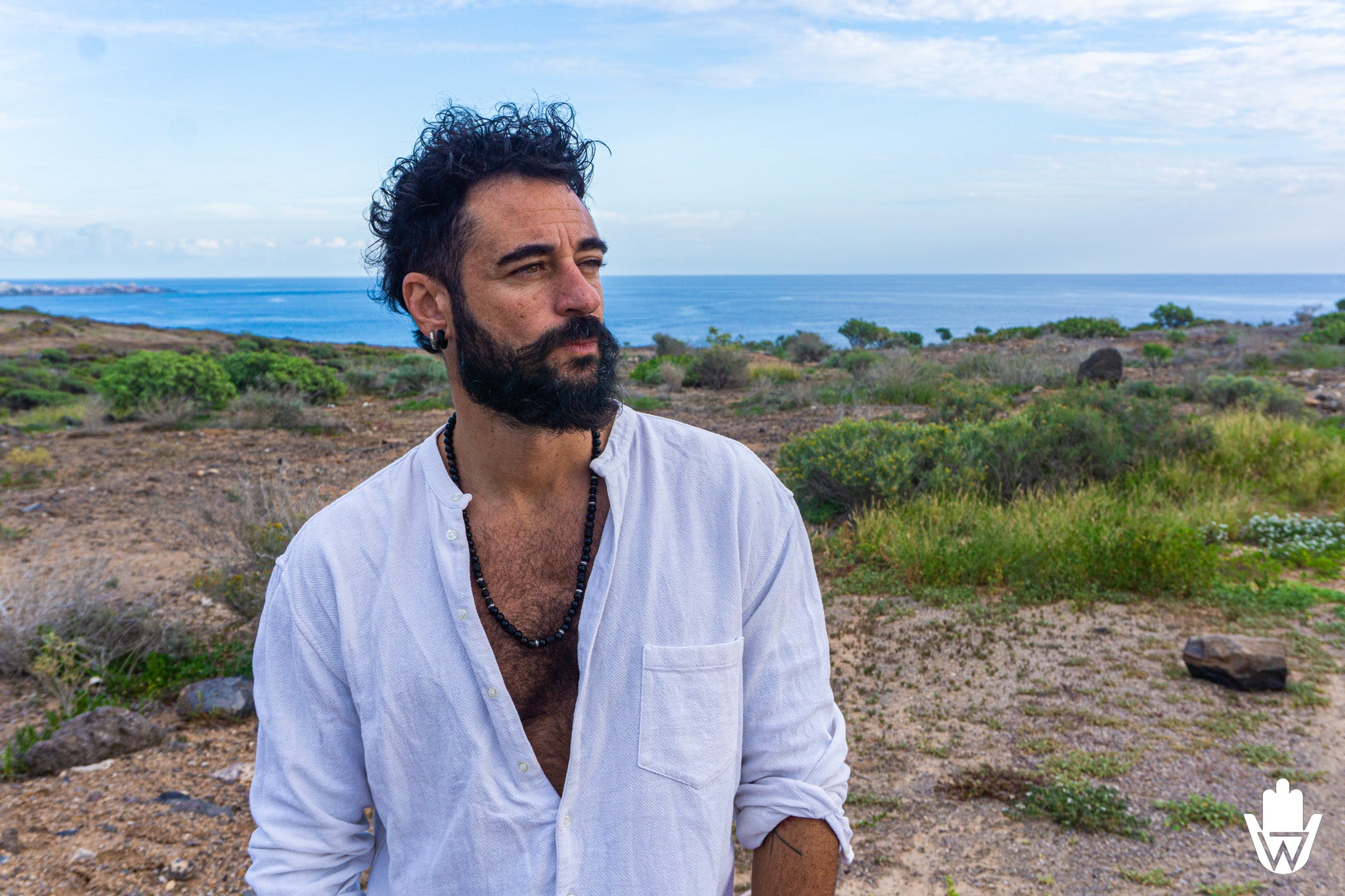 A man with a beard and curly hair wearing an open white button-down shirt stands outdoors in a dry, scrub-covered landscape with the ocean and blue sky in the background.